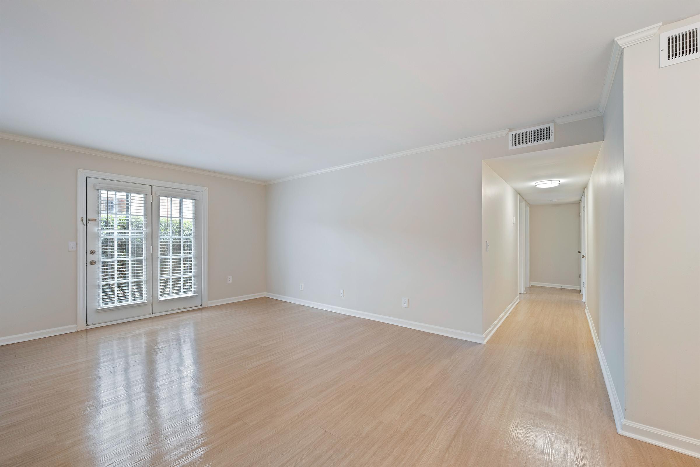 A spacious, empty living room with light-colored walls and wooden flooring. Large windows with white shutters let in natural light. A hallway is visible on the right side, leading to other rooms. The overall ambiance is bright and airy, suitable for various interior design possibilities.