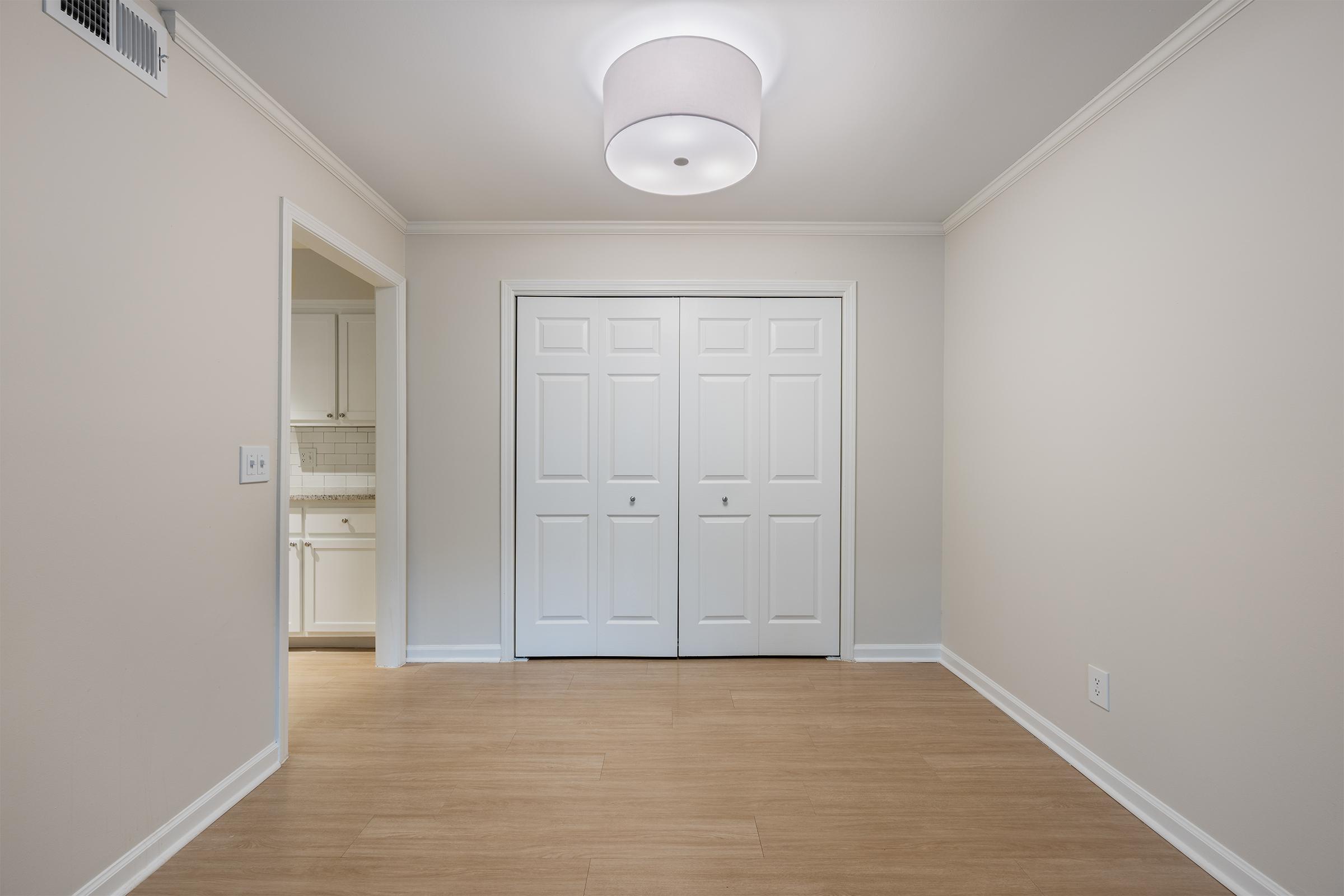 An empty room with light-colored walls and a smooth, light wood floor. It features a modern ceiling light fixture and a double door closet on one side. In the background, a partially visible kitchen area can be seen through an open doorway. The space is bright and uncluttered, suggesting a fresh, inviting atmosphere.