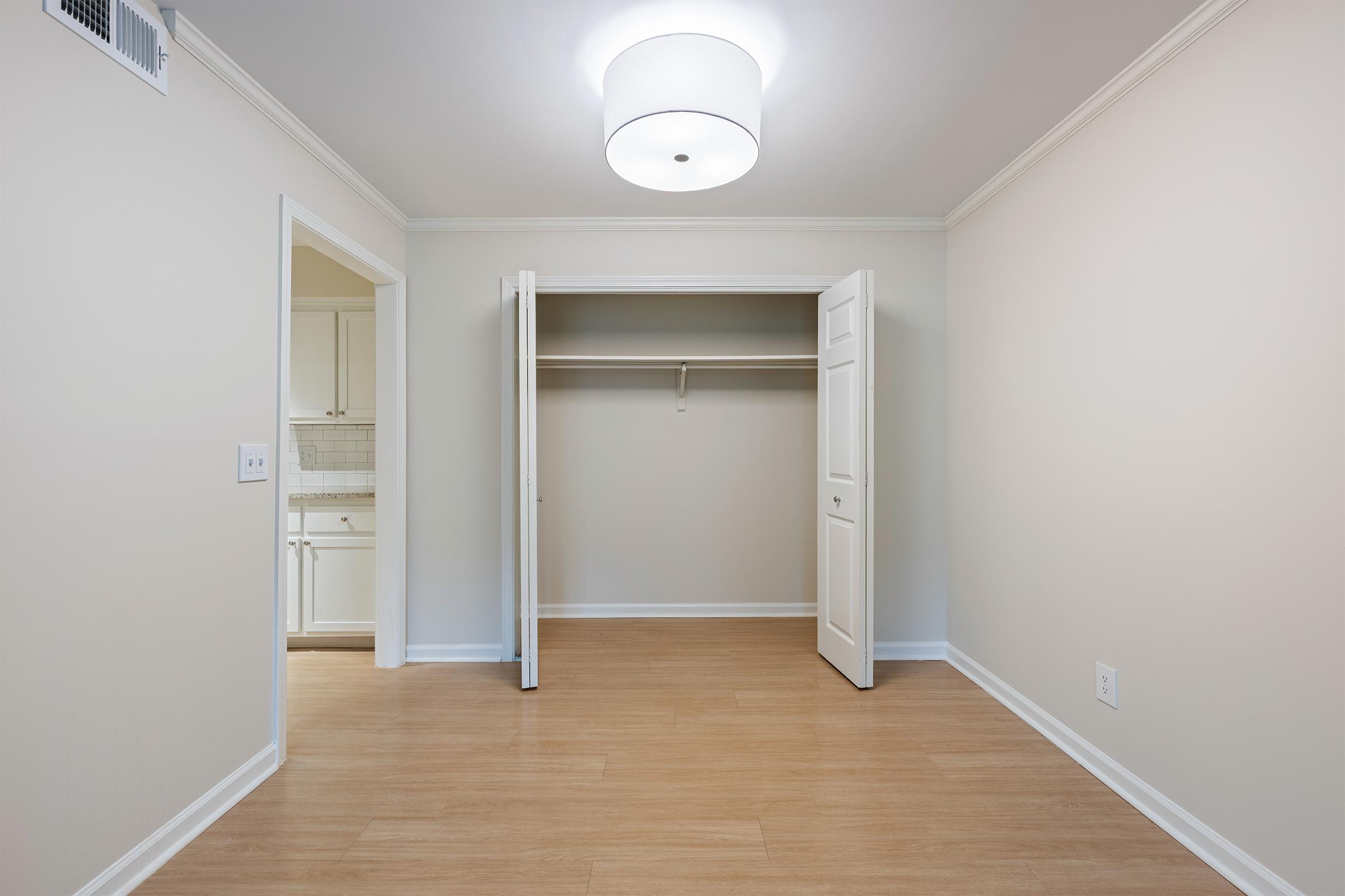 An empty room with light-colored walls and a ceiling light fixture. There is a closet with open double doors, showcasing a simple, uncluttered space. The flooring is light wood, and a doorway leads to another room, likely a kitchen. The overall ambiance is bright and minimalistic.
