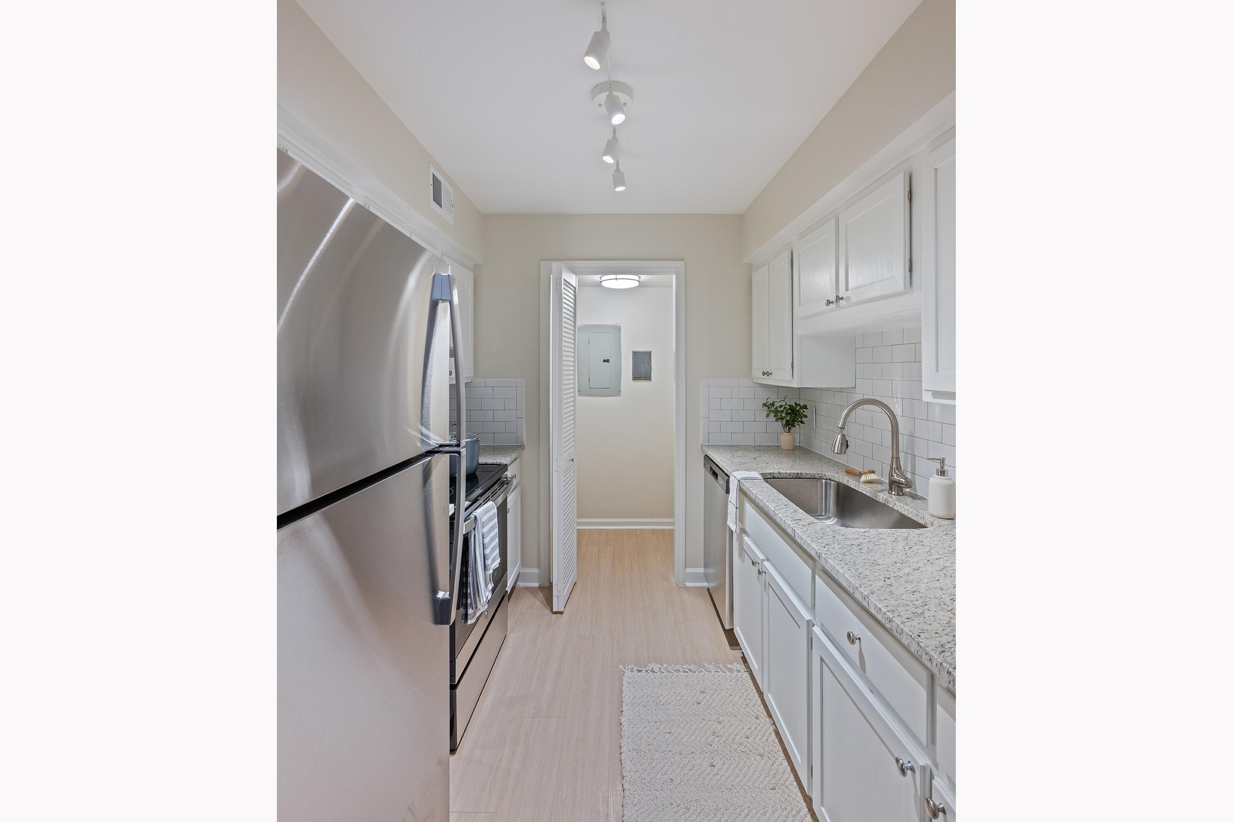 Modern kitchen featuring stainless steel appliances, including a refrigerator and sink with a granite countertop. The space has white cabinetry, tiled backsplash, and a doorway leading to another room, with soft lighting from track fixtures and warm beige walls. A small plant adds a touch of greenery.