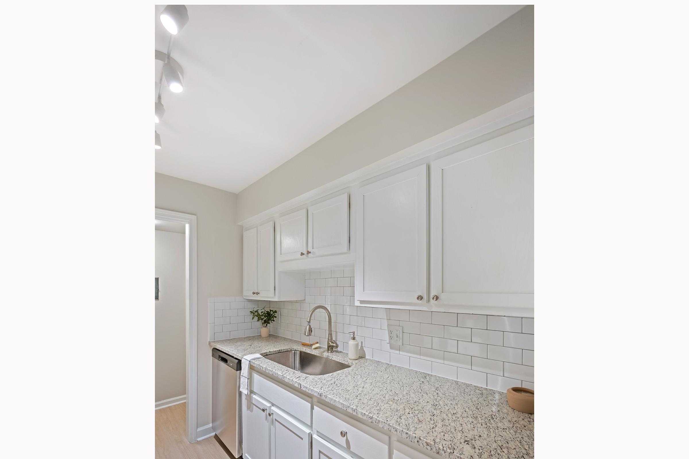 A modern kitchen featuring white cabinets, a granite countertop, and a stainless steel sink. The backsplash consists of white subway tiles, and there's a small potted plant on the counter. The space is well-lit with recessed lighting, giving it a bright and airy feel.