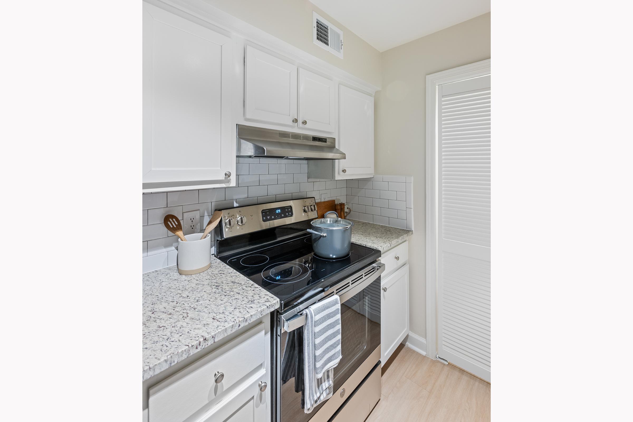 Modern kitchen featuring white cabinets, a silver stove with an oven, and a granite countertop. A pot is placed on the stove, along with kitchen utensils in a holder. The backsplash consists of white subway tiles, and there is a door leading to a pantry or storage area. Bright and clean design.