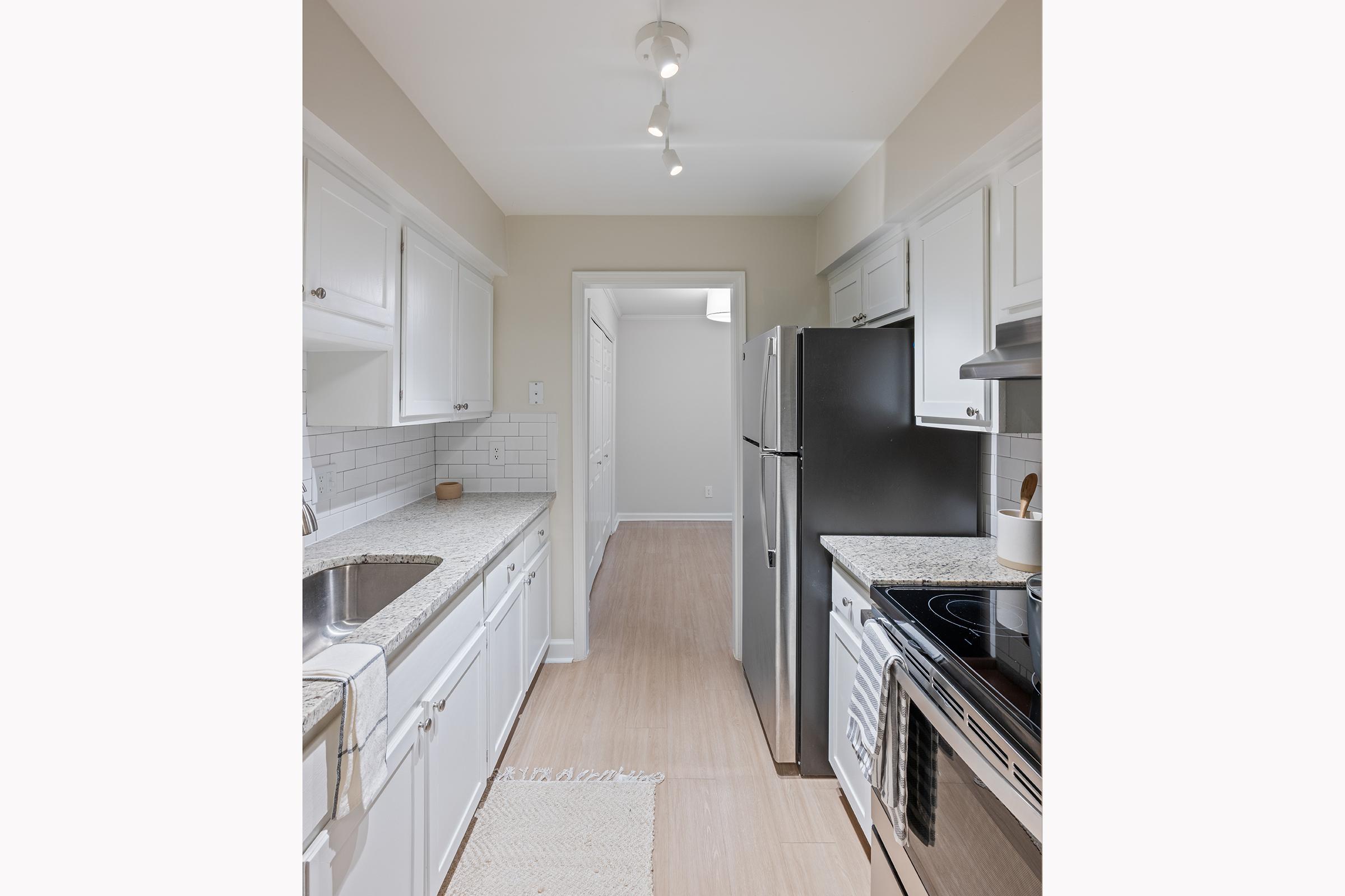 A modern kitchen featuring white cabinets, a double sink, granite countertops, and stainless steel appliances including a refrigerator and stove. The space is well-lit with a ceiling fixture and has light-colored flooring, leading to an adjoining room.
