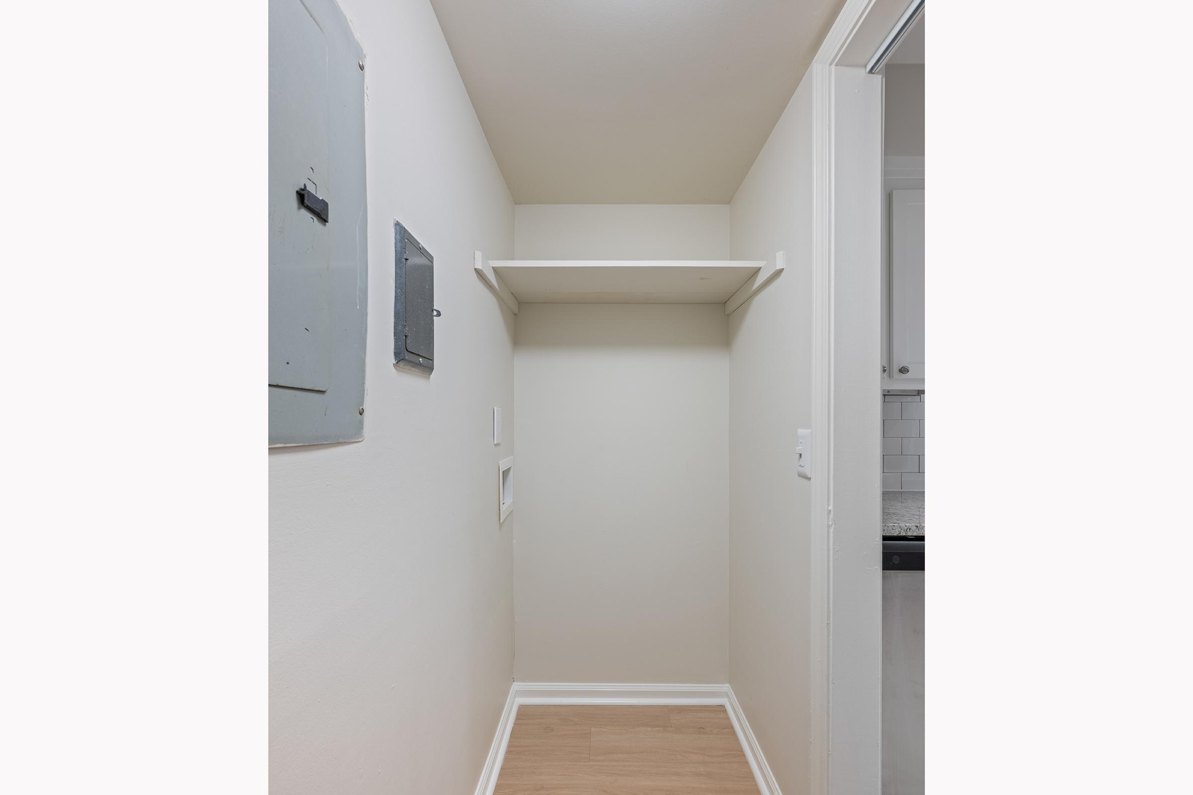 A small, empty closet with a single shelf against a white wall. There is an electrical panel on the left side and a smooth, light-colored floor. The closet is well-lit and has a minimalistic design, with no visible items or decorations.