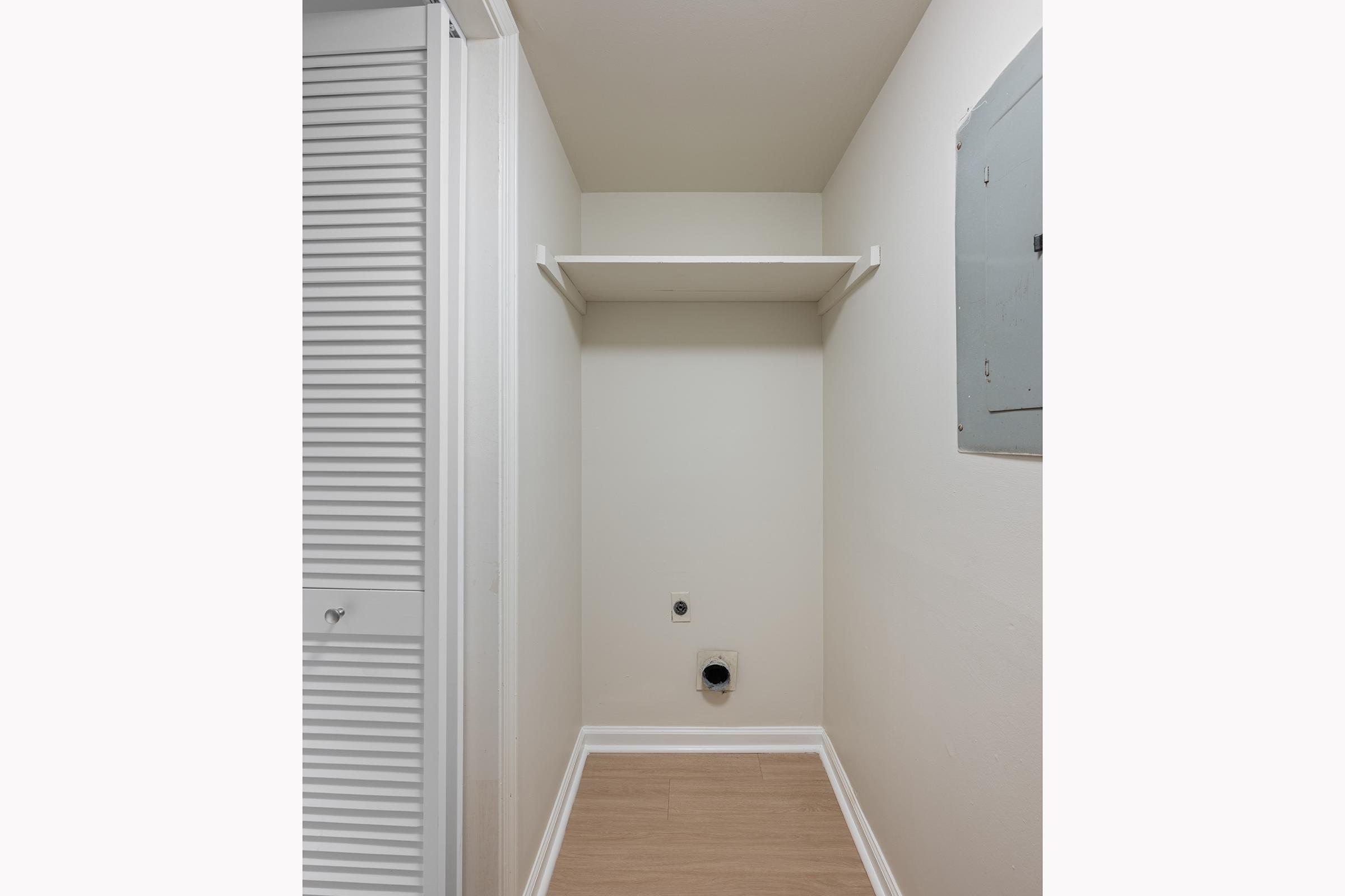 A small, empty laundry room with light-colored walls and a shelf mounted on the back wall. The floor is a light wood laminate. There is a white closet door on the left and an electrical panel on the right, with plumbing connections visible on the floor. The space is well-lit and clean.