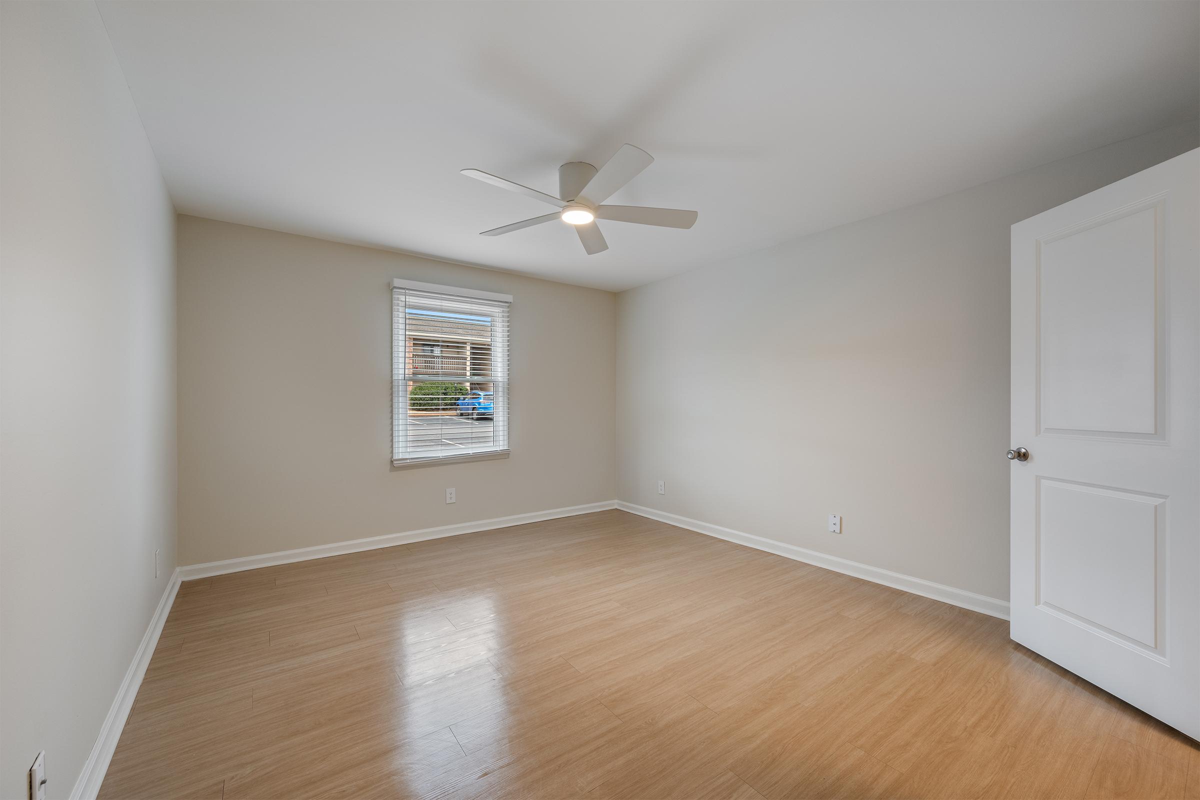 A clean, empty room featuring light-colored walls and a hardwood floor. A window with blinds allows natural light to enter, and a modern ceiling fan is installed. A white door is visible on the right side, completing the simple, minimalist design of the space.