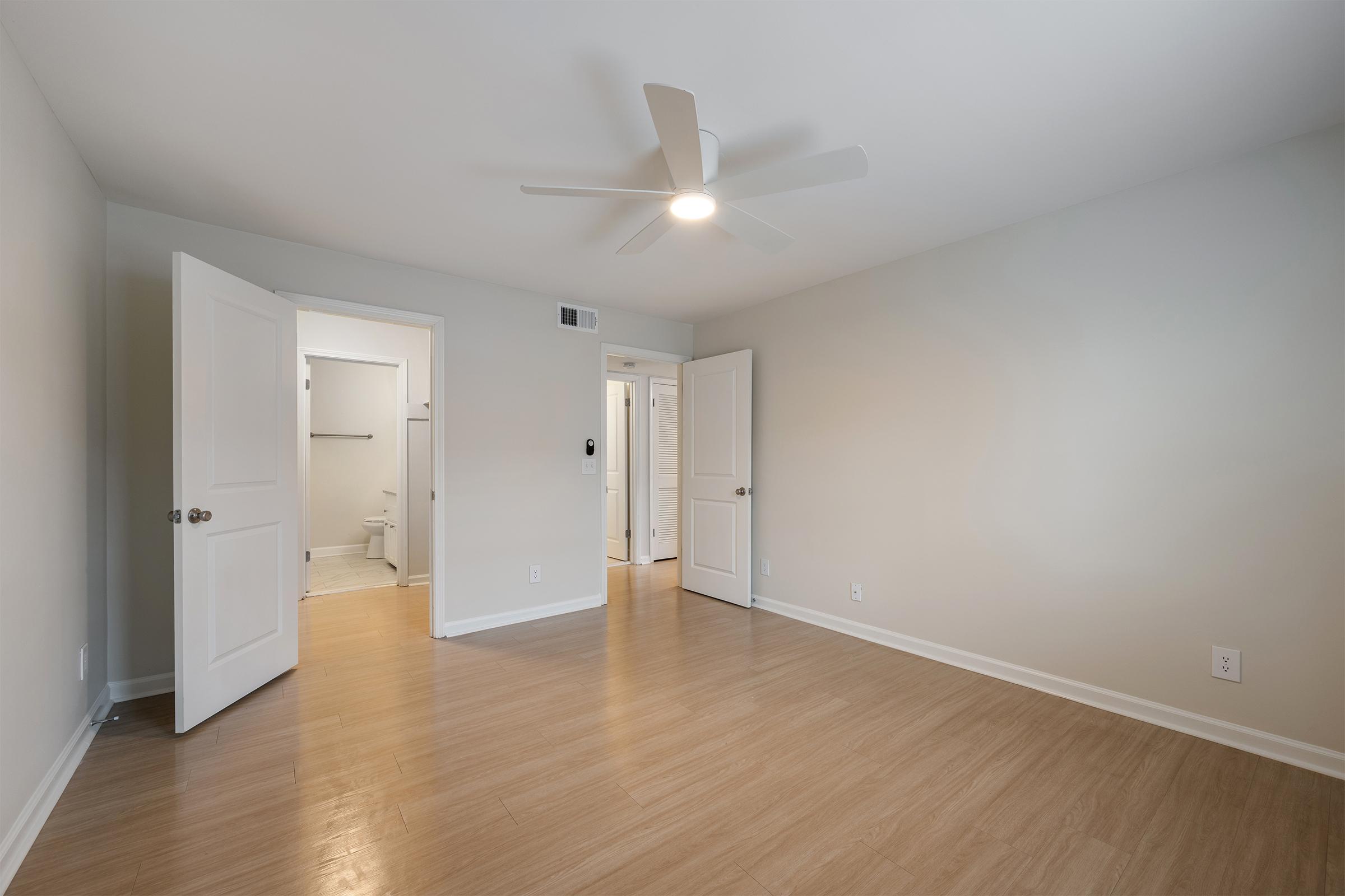 A bright and spacious room with light beige walls and hardwood flooring. There are two open doors leading to adjacent spaces; one appears to be a bathroom. A ceiling fan is visible above, providing additional light and ventilation. The overall ambiance is clean and uncluttered.