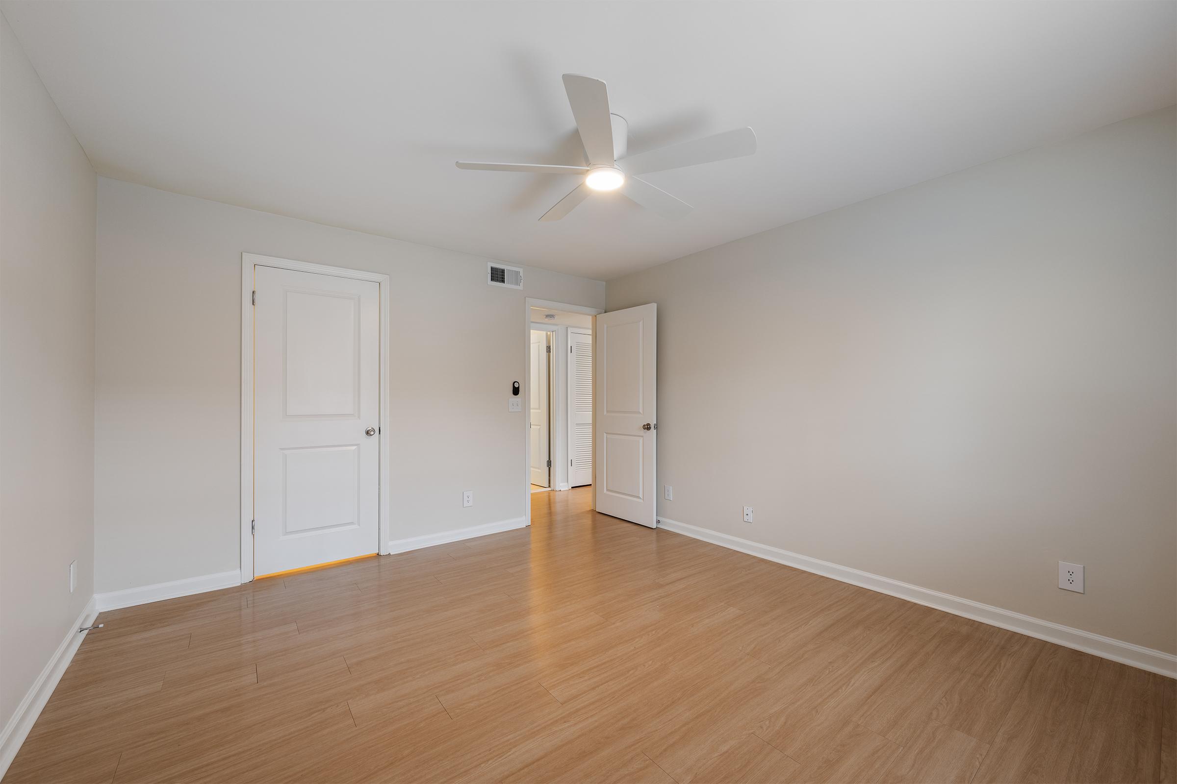 Empty room with light-colored walls and a ceiling fan. Features a wooden floor, two doors on opposite walls, and minimal furnishings. Soft natural light illuminates the space, creating a clean and spacious look.