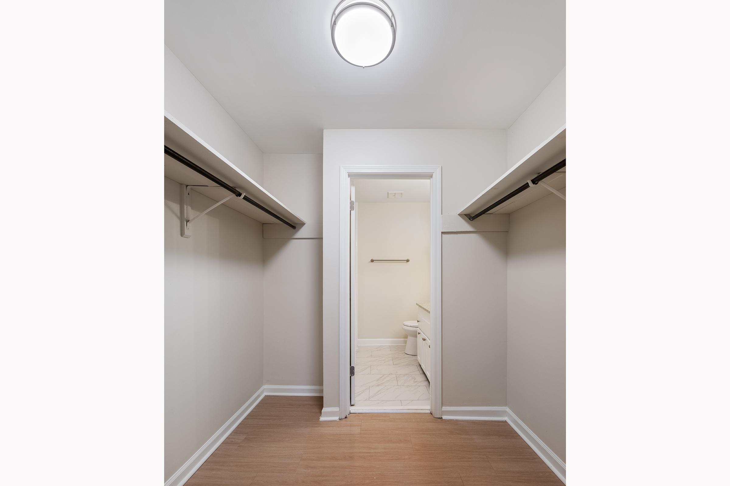 A spacious, empty walk-in closet featuring two hanging rods on either side, light-colored walls, and a laminate floor. A doorway at the back leads to a bathroom with a toilet and a towel bar visible. The ceiling has a round light fixture providing illumination.
