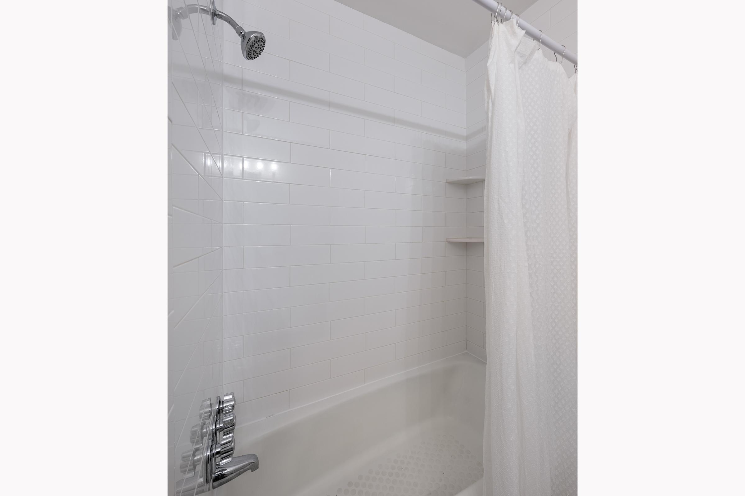 A clean, modern bathroom featuring a white tiled shower area with a chrome showerhead. The space includes a bathtub and a sheer white shower curtain, along with minimalist shelves on the wall. The design is bright and contemporary, emphasizing simplicity and functionality.
