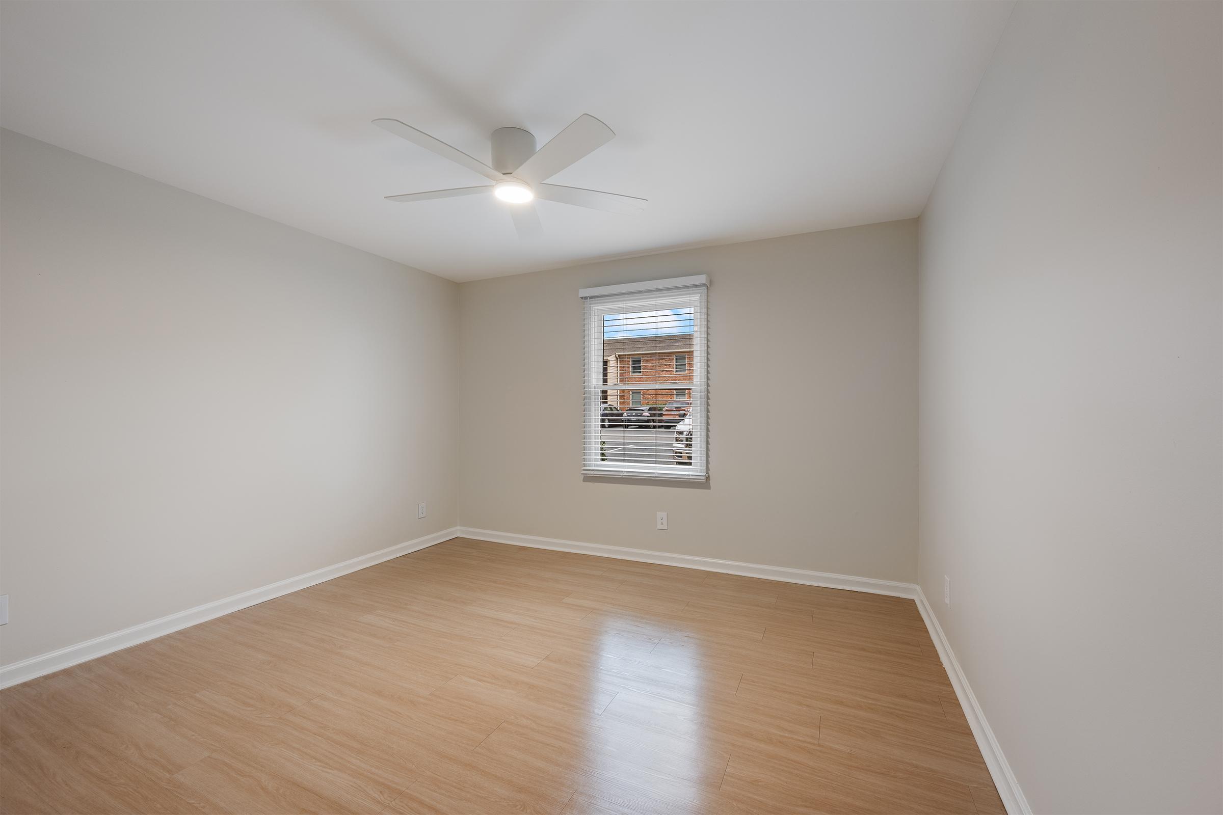A spacious, empty room with light-colored walls and a wooden floor. A ceiling fan is mounted in the center, and there is a window with blinds allowing natural light to enter. The view outside the window shows a brick building. The room appears clean and well-maintained.