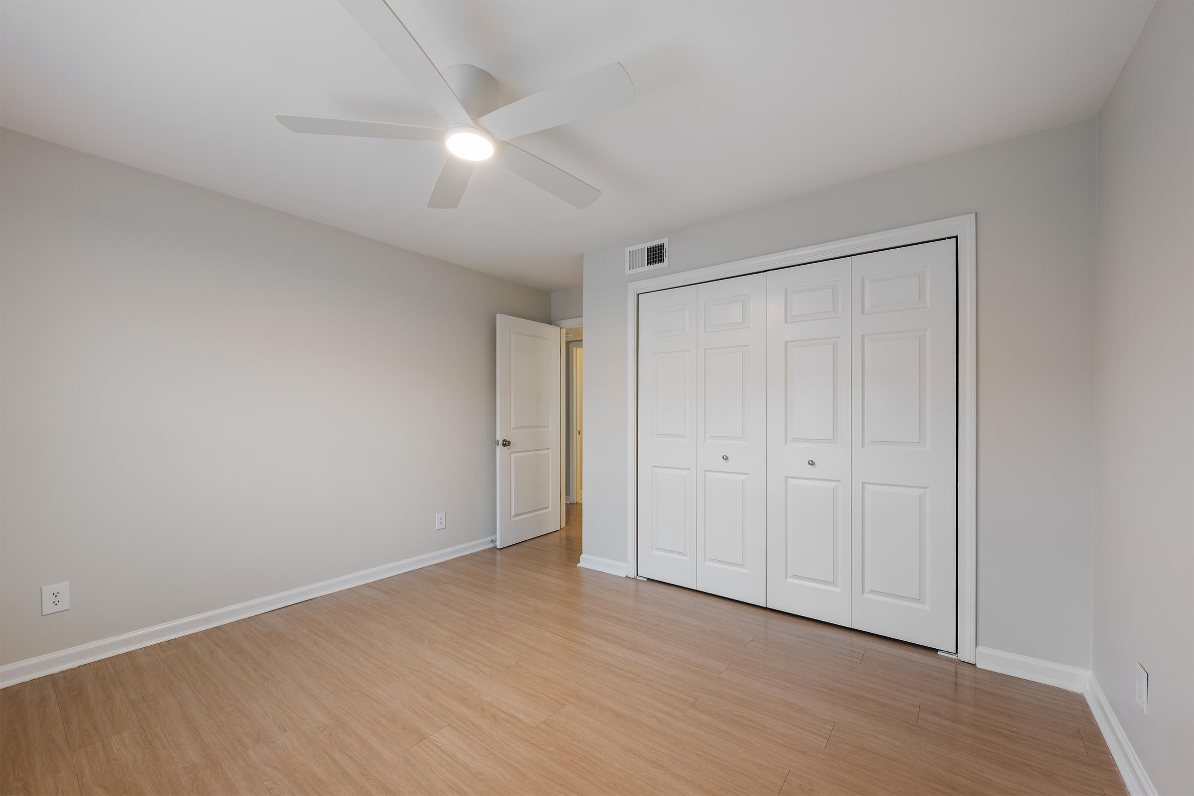 A spacious, empty room featuring light-colored walls, a ceiling fan, and laminate flooring. There's a white double door closet on one side and an open door leading to another room. The overall ambiance is bright and clean, suitable for various uses.