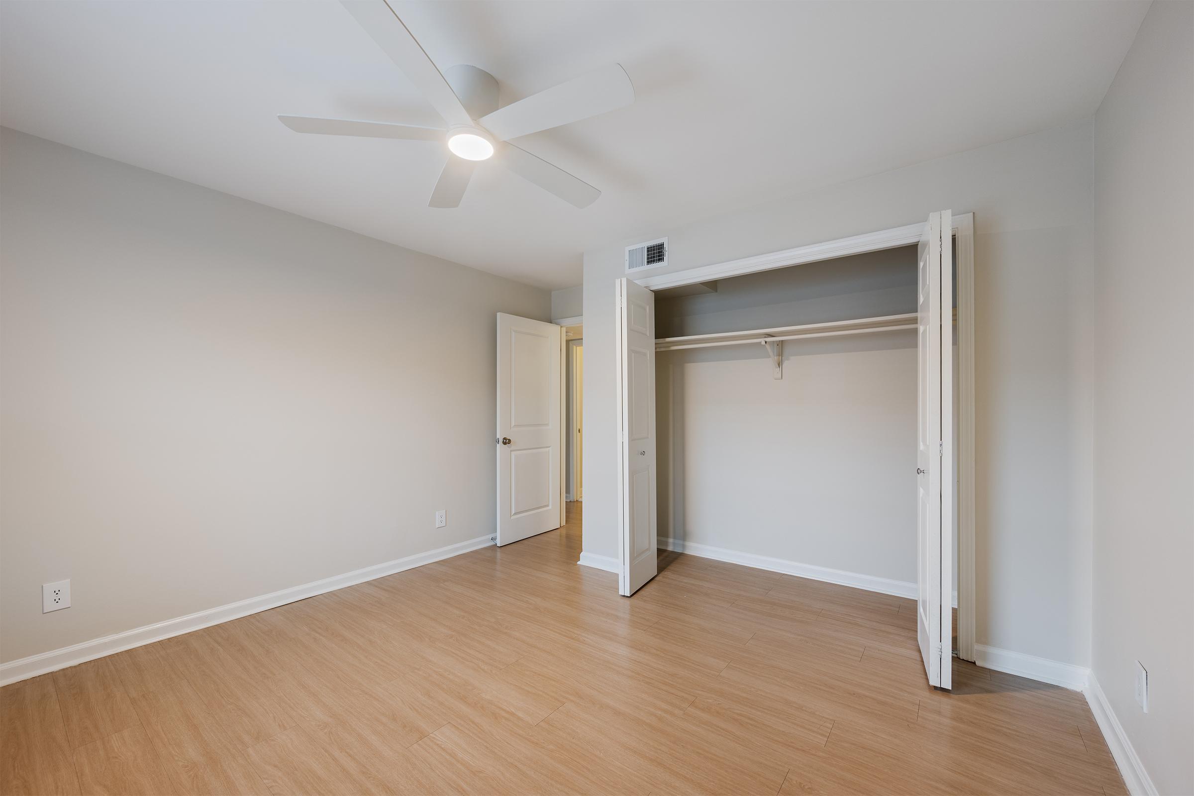 A well-lit, empty room featuring pale walls and a ceiling fan. It has light wood flooring and a closet with open double doors, revealing an unadorned interior. A doorway leads to another room, suggesting a spacious layout.