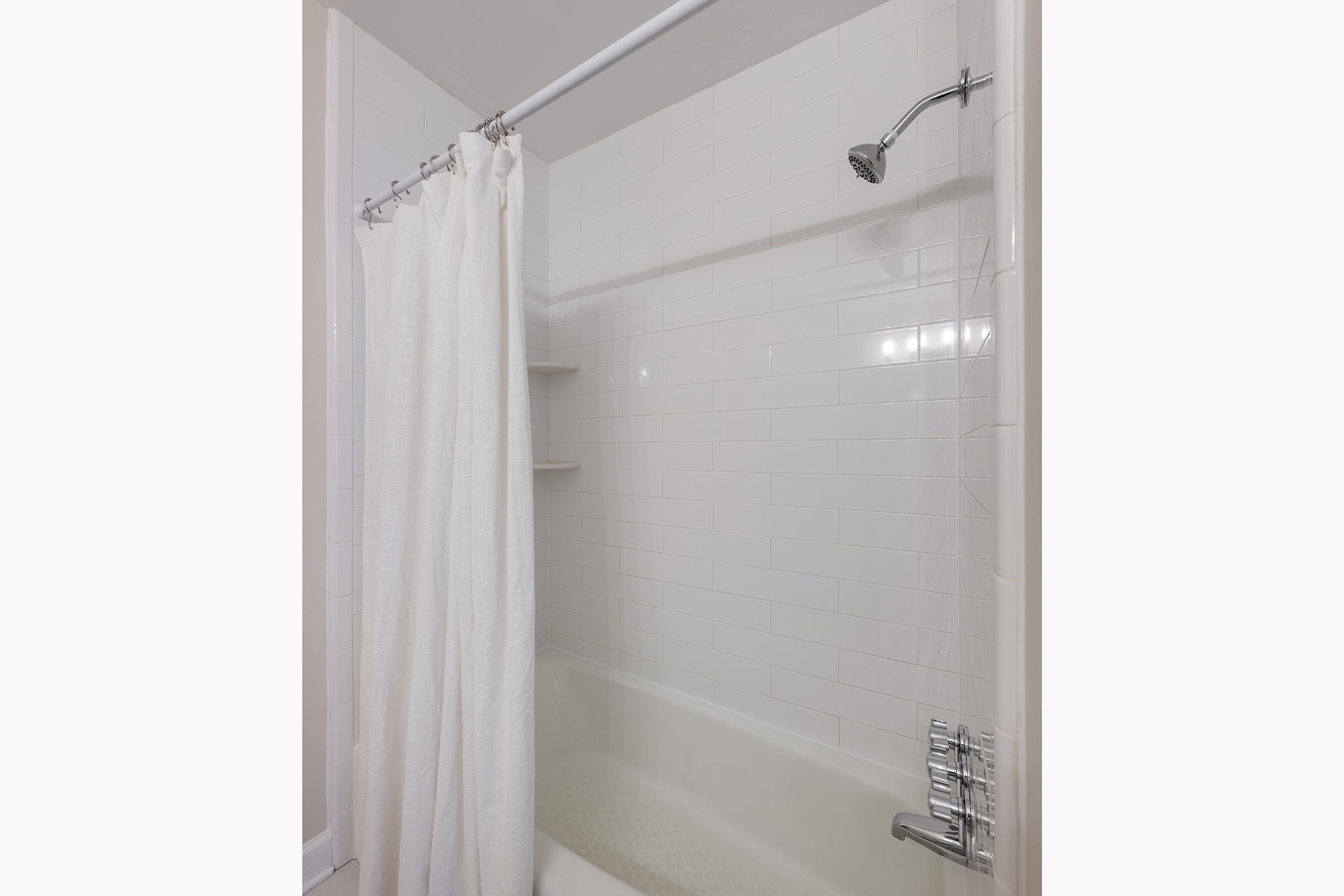 A clean bathroom shower area featuring a white tub with a modern faucet, surrounded by white tiled walls. A white shower curtain hangs from a chrome rod, and there are floating shelves on one side for toiletries. The overall aesthetic is simple and contemporary.