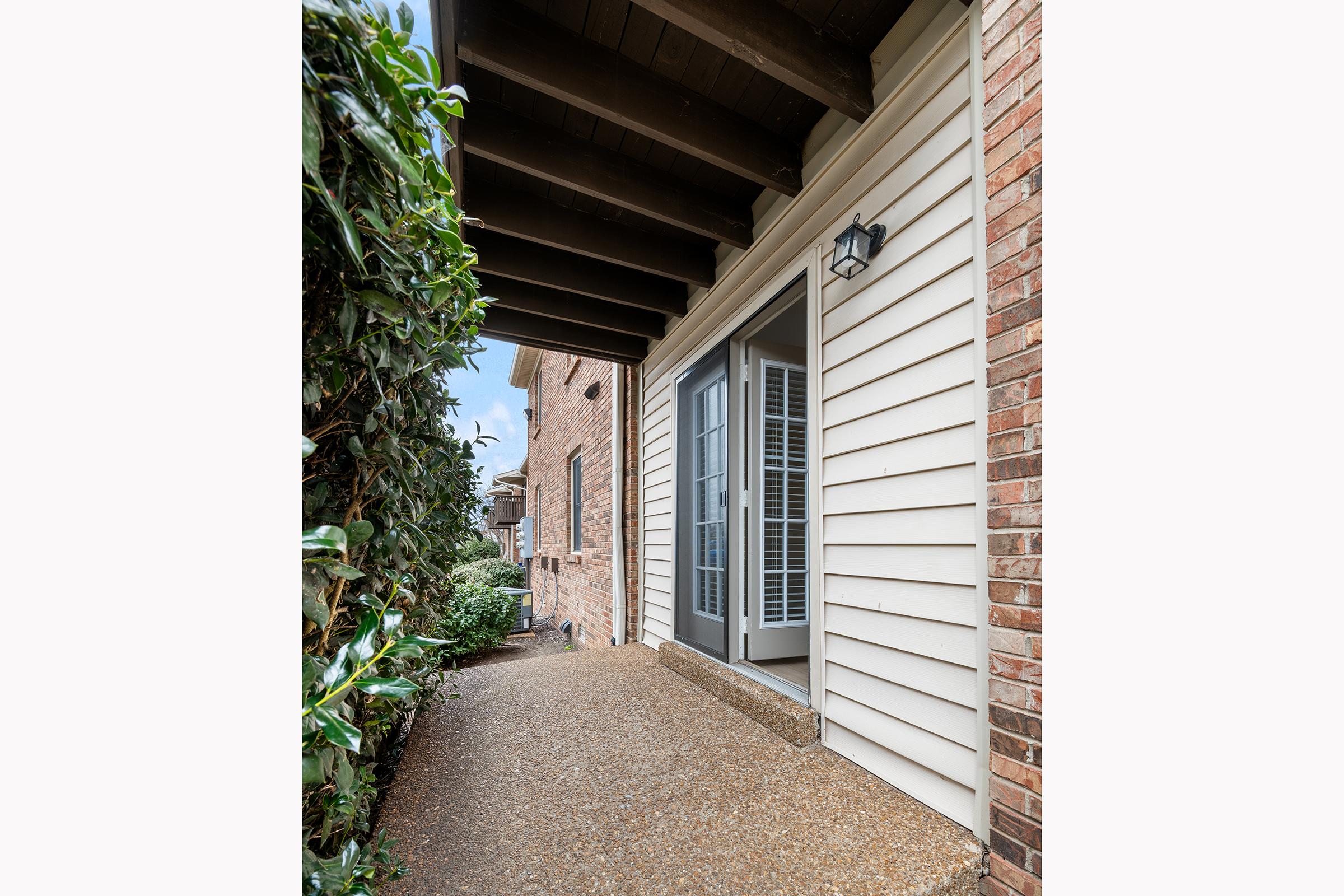 A narrow pathway alongside a building, featuring a light-colored wooden exterior and a door with windows. Lush green foliage is visible on one side, while the pathway is made of smooth gravel. The overhead beams create a shaded area, and the scene is set under an overcast sky.