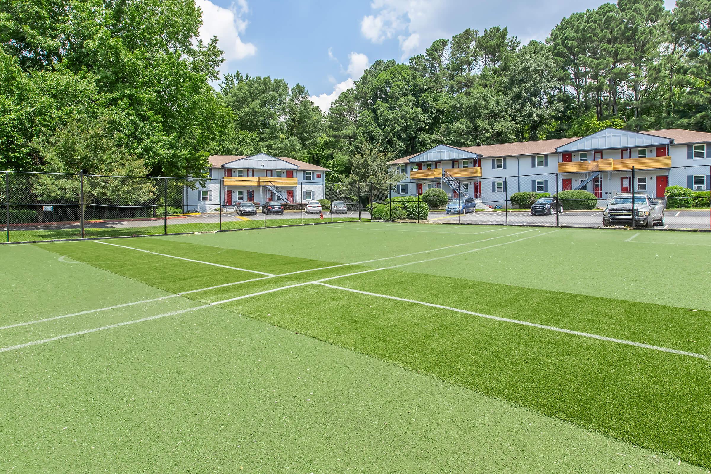 A view of a well-maintained artificial turf tennis court with white lines crossing in the foreground. In the background, there are several residential buildings with colorful exteriors surrounded by trees and parking spaces. The sky is partly cloudy, creating a bright and inviting atmosphere.