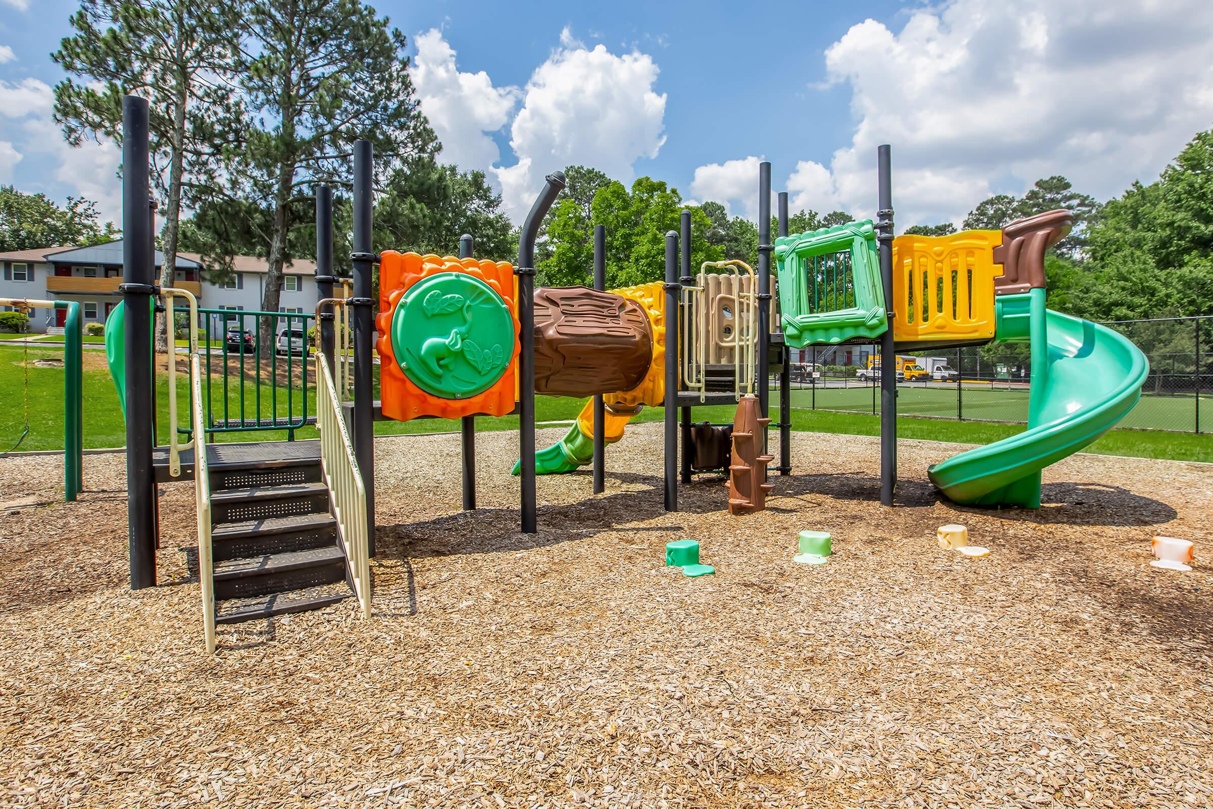 Colorful playground equipment featuring a climbing structure, a slide, and various interactive panels in a park setting. The ground is covered with wood chips, and there are trees and residential buildings in the background under a clear blue sky with fluffy white clouds.