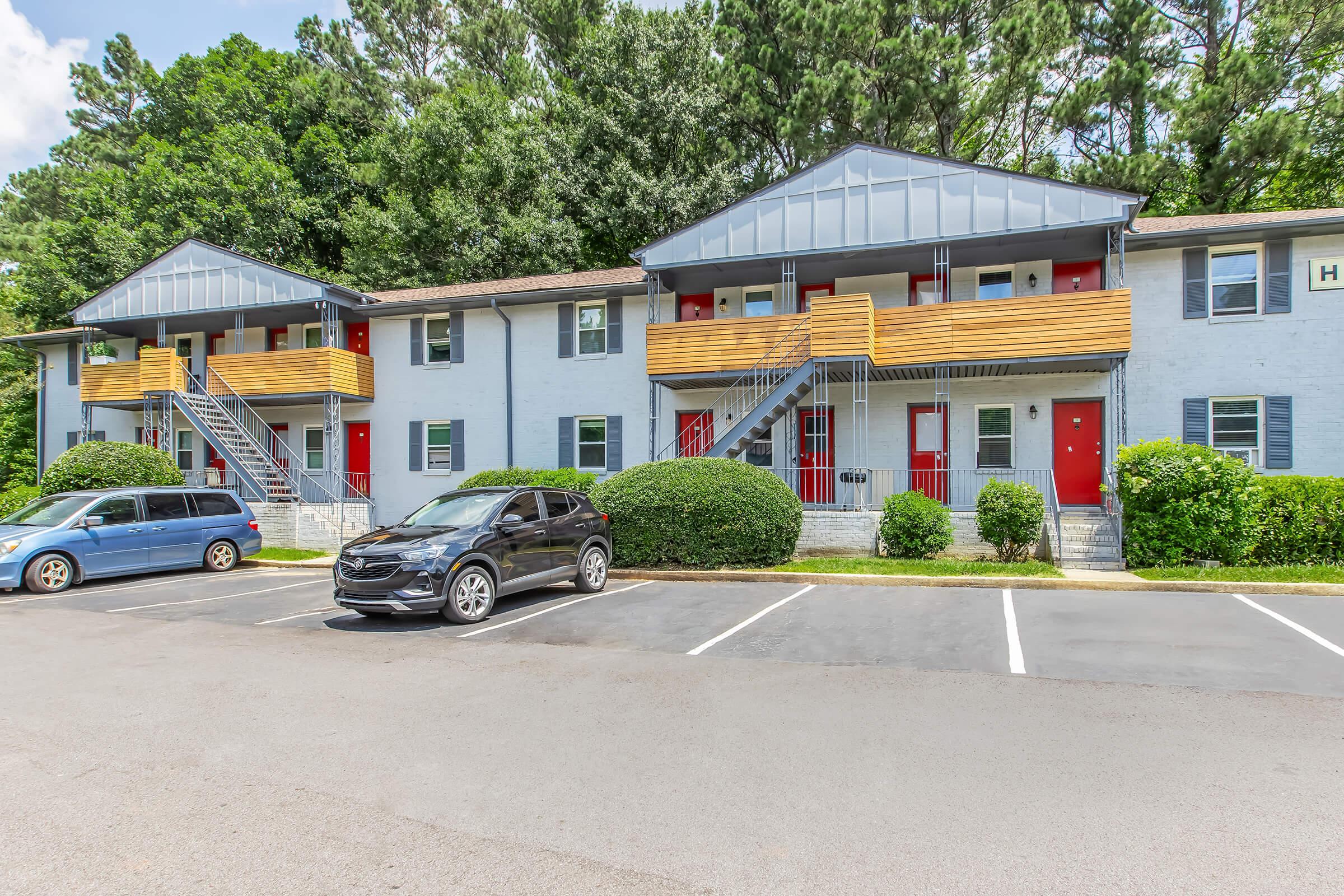 A view of a multi-unit residential building with two stories. The building features a light gray exterior, wooden balconies with red doors, and well-maintained shrubs. Two parked cars are visible in the foreground, along with a paved parking lot and surrounding greenery.