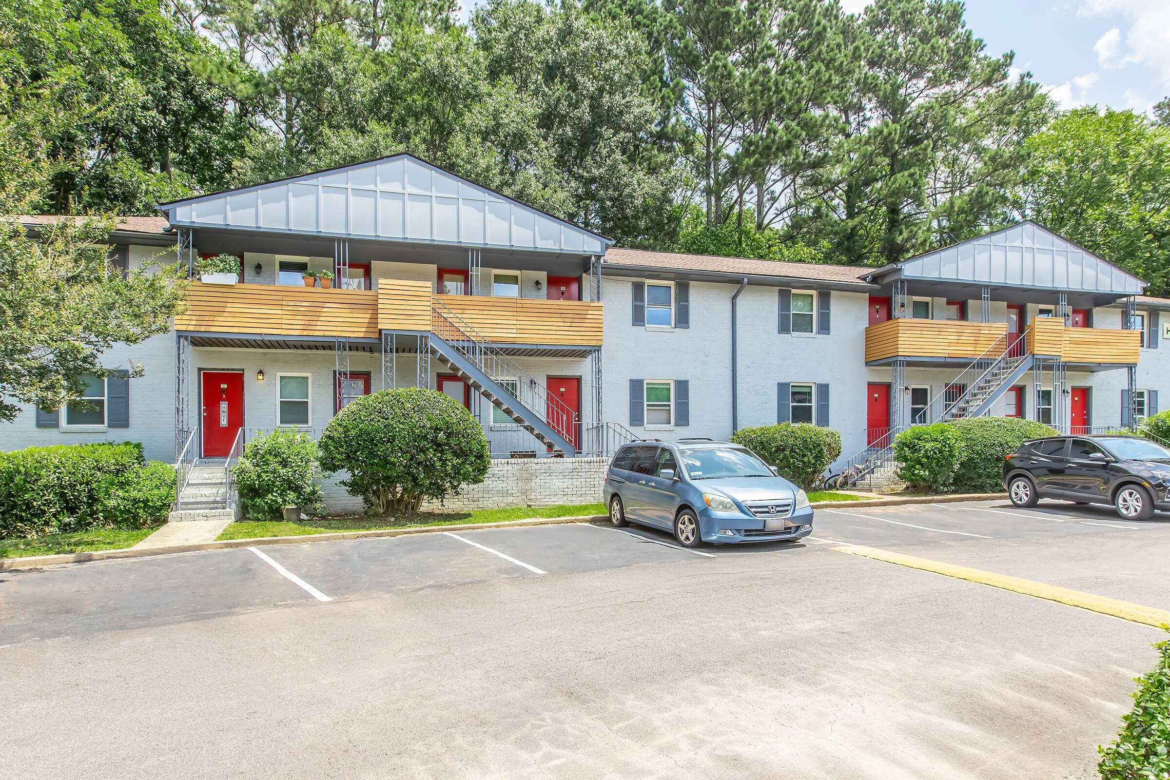 A view of a two-story apartment building with three units featuring red doors and wooden balconies. The building is surrounded by greenery, with a paved parking area in front. Two cars are parked in the lot, and trees can be seen in the background under a blue sky.
