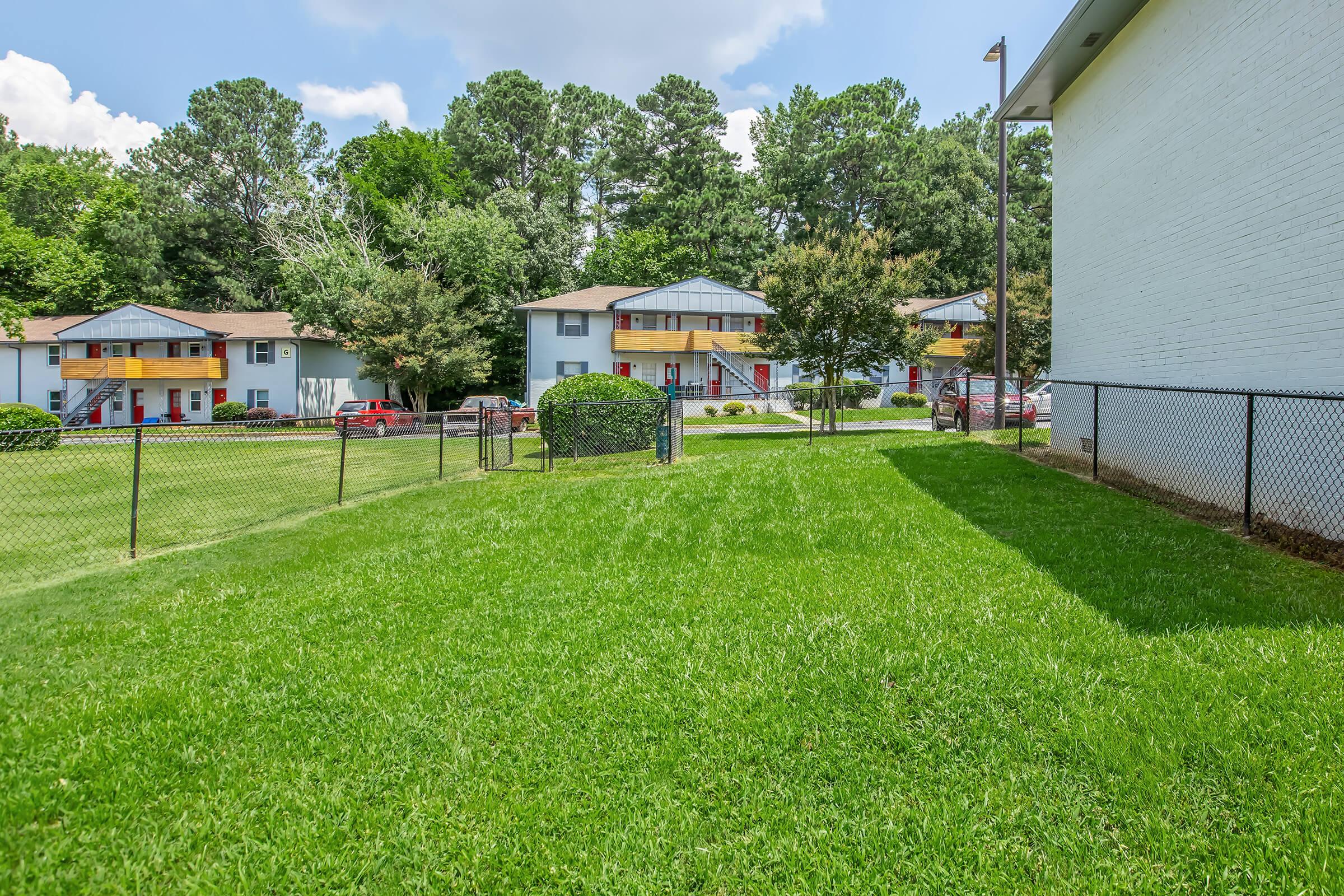 A grassy area enclosed by a fence, with several multi-unit buildings in the background. The buildings feature colorful red doors and are surrounded by lush trees. A clear blue sky and fluffy clouds above contribute to a pleasant, sunny atmosphere.