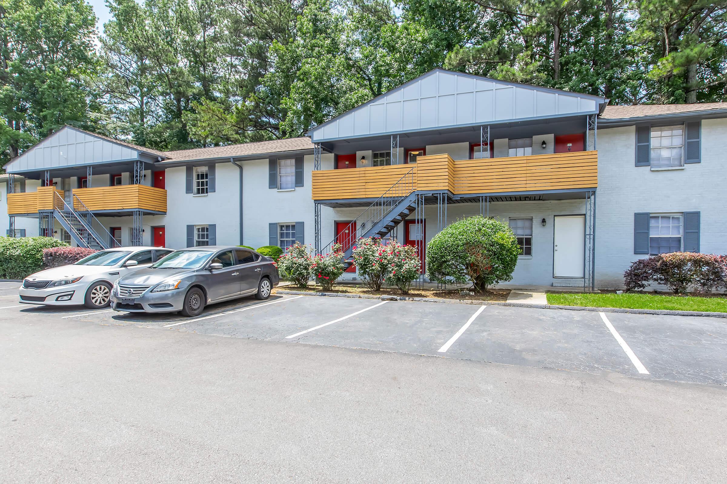 A view of a two-story apartment complex with light blue walls and wooden balconies. The building has red doors and is surrounded by greenery, including shrubs and trees. A few parked cars are visible in the front parking lot, and the area appears well-maintained and inviting.