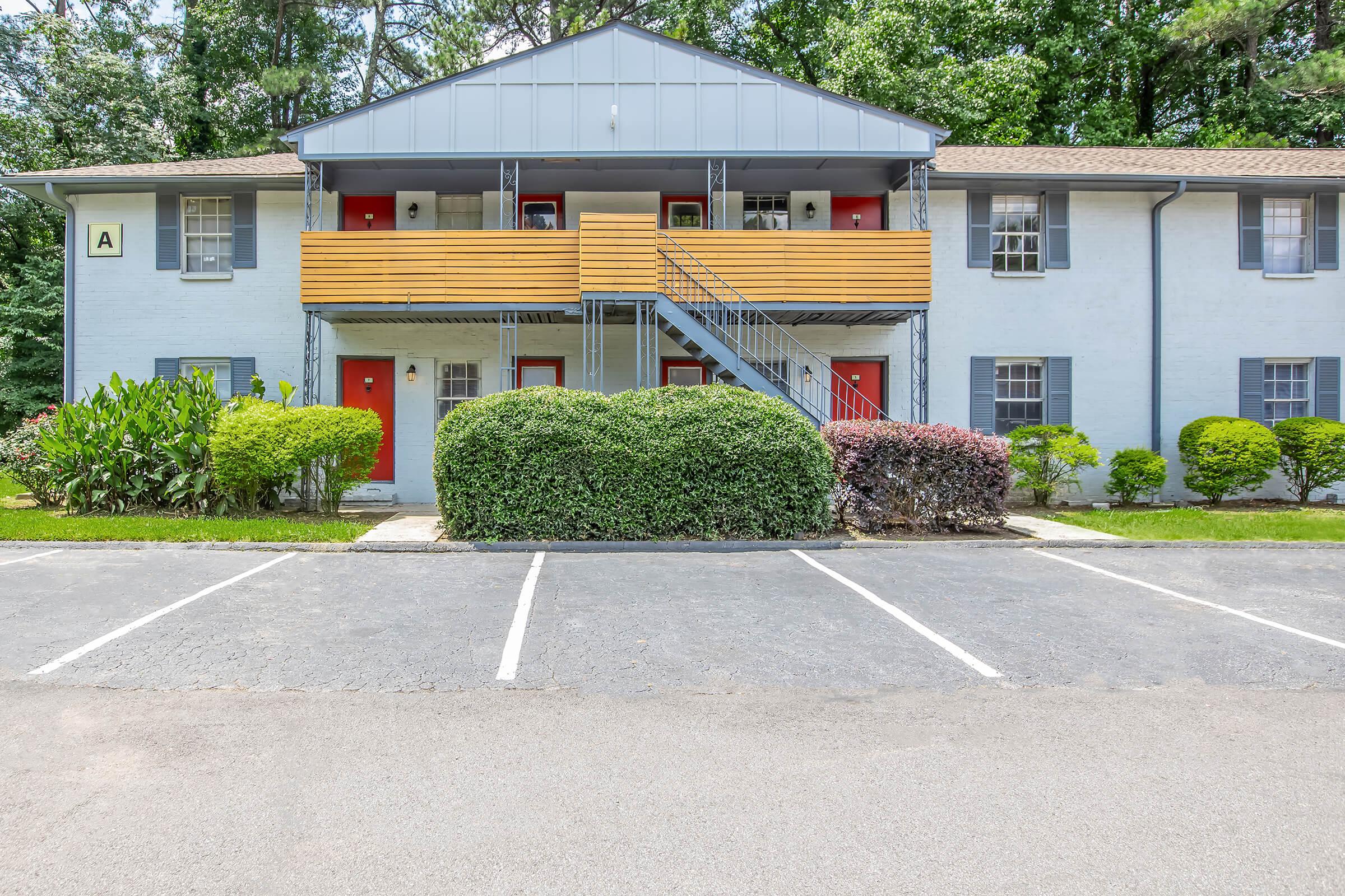 A two-story apartment building with a light blue exterior and red doors, featuring wooden balconies on the upper level. The entrance is flanked by neatly trimmed shrubs and small hedges. A parking lot with several empty spaces is visible in the foreground, surrounded by greenery and trees in the background.