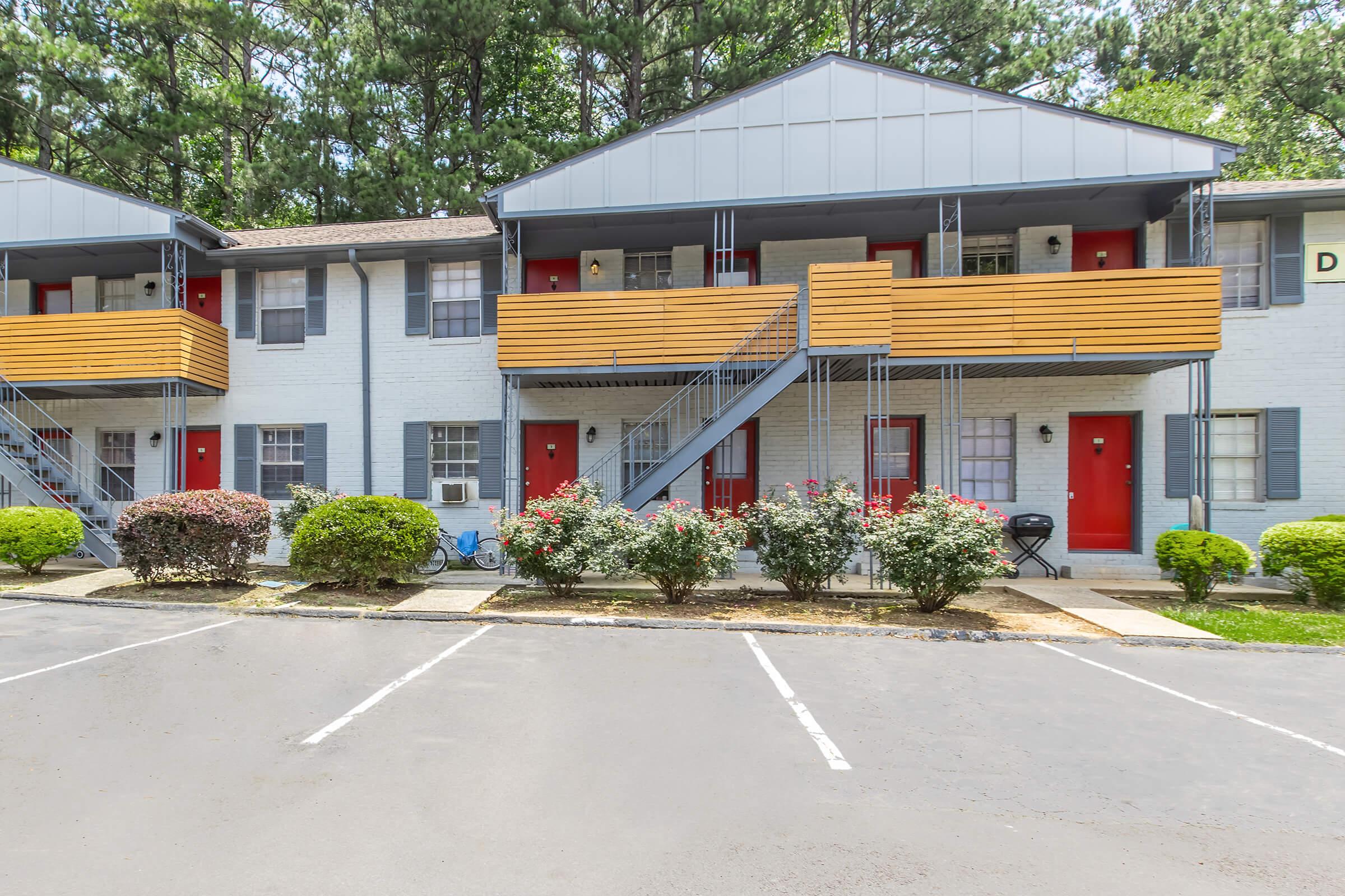 A two-story apartment building with light gray brick walls and wooden balconies. The first floor has red front doors complemented by neatly trimmed bushes and small trees. A parking lot is visible in front, and the area is surrounded by tall trees in the background, creating a tranquil setting.