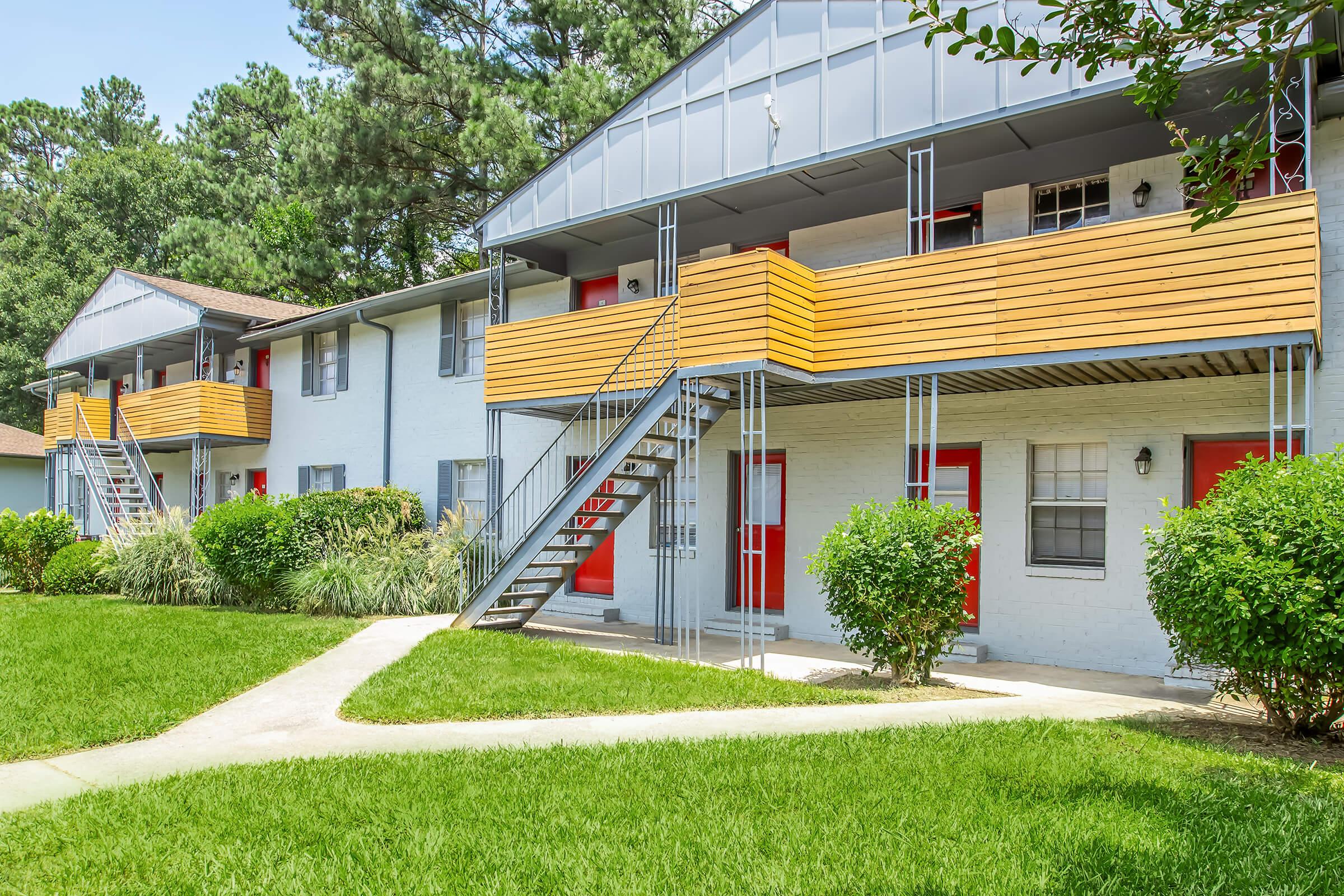 Two two-story apartment buildings with wooden balconies and red doors, surrounded by well-manicured lawns and hedges. A concrete pathway runs through the grass in front of the buildings, and tall trees are visible in the background under a clear blue sky.