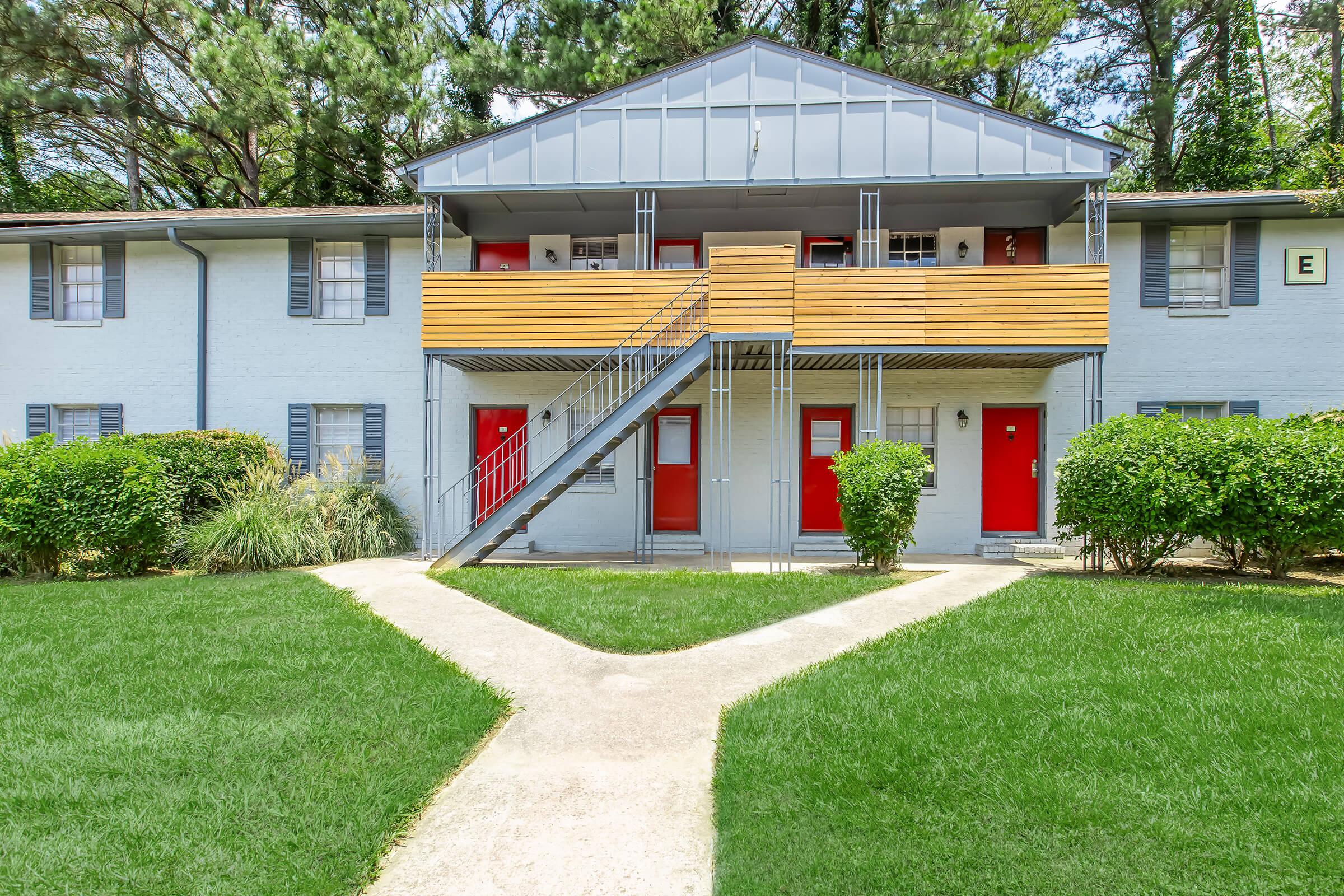 A two-story apartment building with blue exterior walls, wooden balconies, and bright red doors. A central walkway leads to the entrance, surrounded by well-maintained green lawns and shrubs. Trees are visible in the background, adding to the serene outdoor setting.