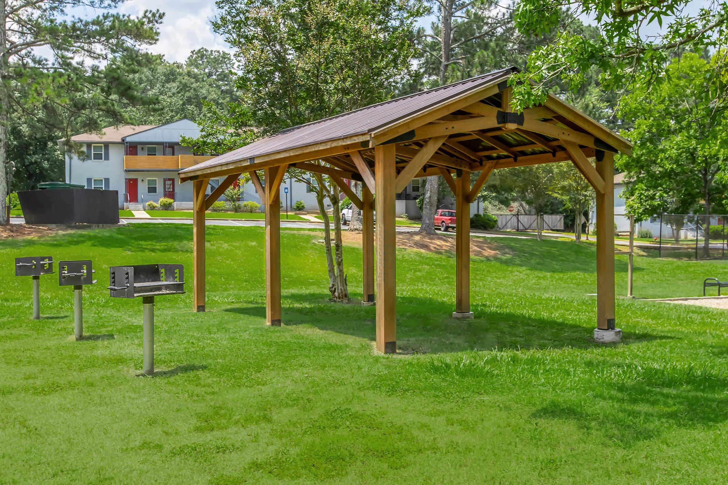 A wooden pavilion with a sloped roof is situated in a green park, surrounded by grass and trees. Nearby, there are several barbecue grills for public use. In the background, there are residential buildings visible, contributing to a community atmosphere.