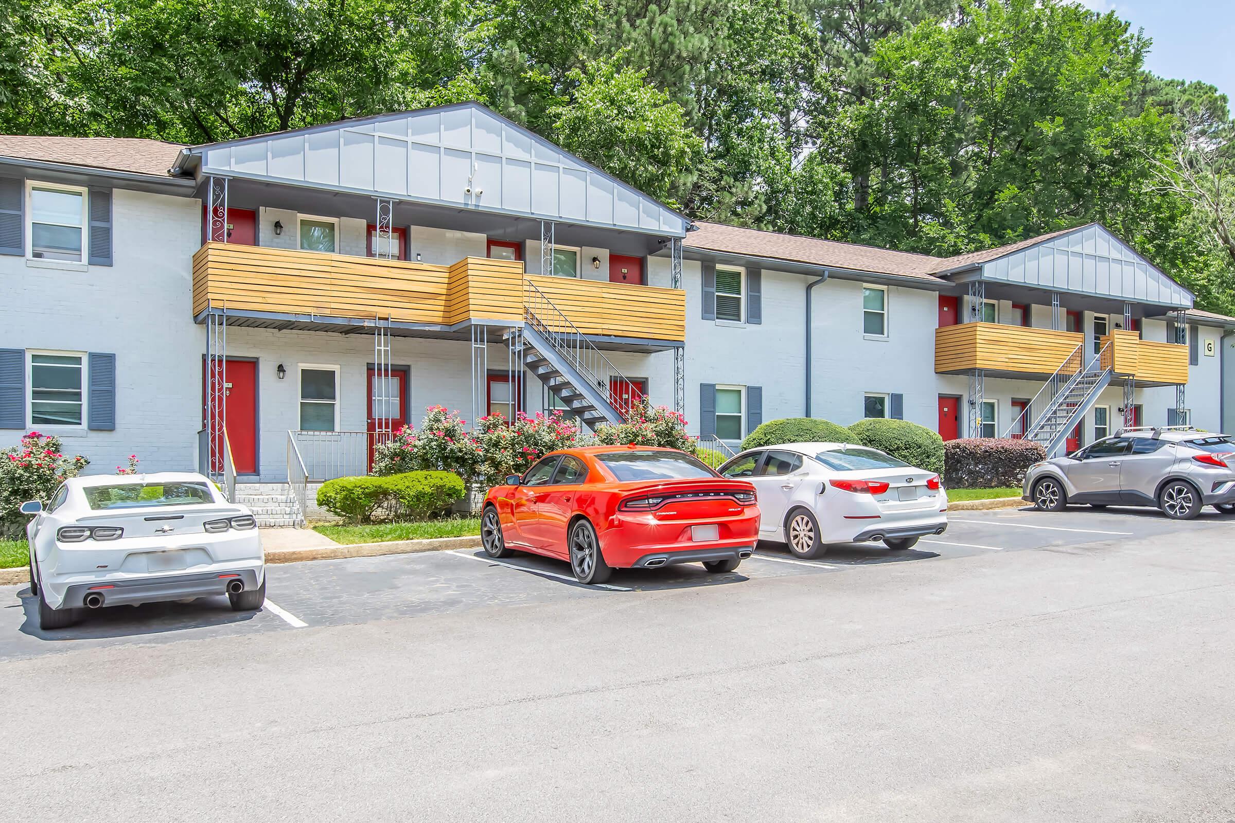 Rows of apartment buildings featuring a mix of colors, with wooden balconies and a well-maintained garden. Several cars parked in front, including a bright red sports sedan, a white SUV, and a silver compact car, surrounded by lush green trees in the background.