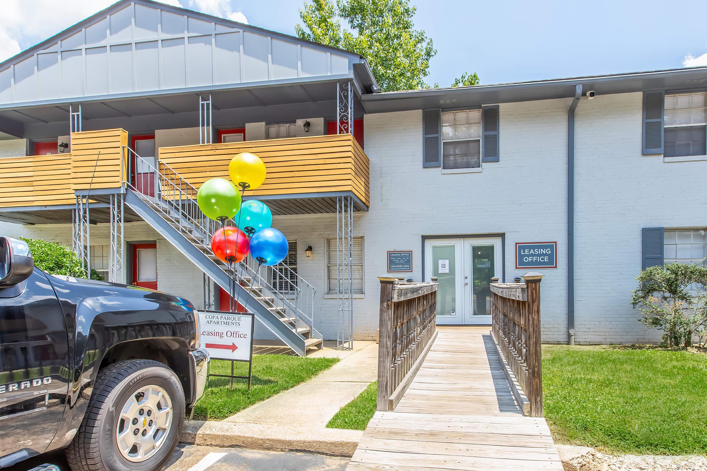 Leasing office of an apartment complex featuring a wooden ramp leading to the entrance. Colorful balloons in blue, red, yellow, and green are attached to a sign post. A black truck is parked in front. The building is two stories with balconies and green landscaping around the walkway.