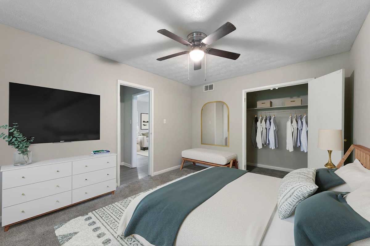 A modern bedroom featuring a large bed with decorative pillows, a wall-mounted TV, and a white dresser. A spacious closet with hanging clothes is visible, along with a cozy bench at the foot of the bed and a ceiling fan above, all set against neutral-colored walls and carpet.