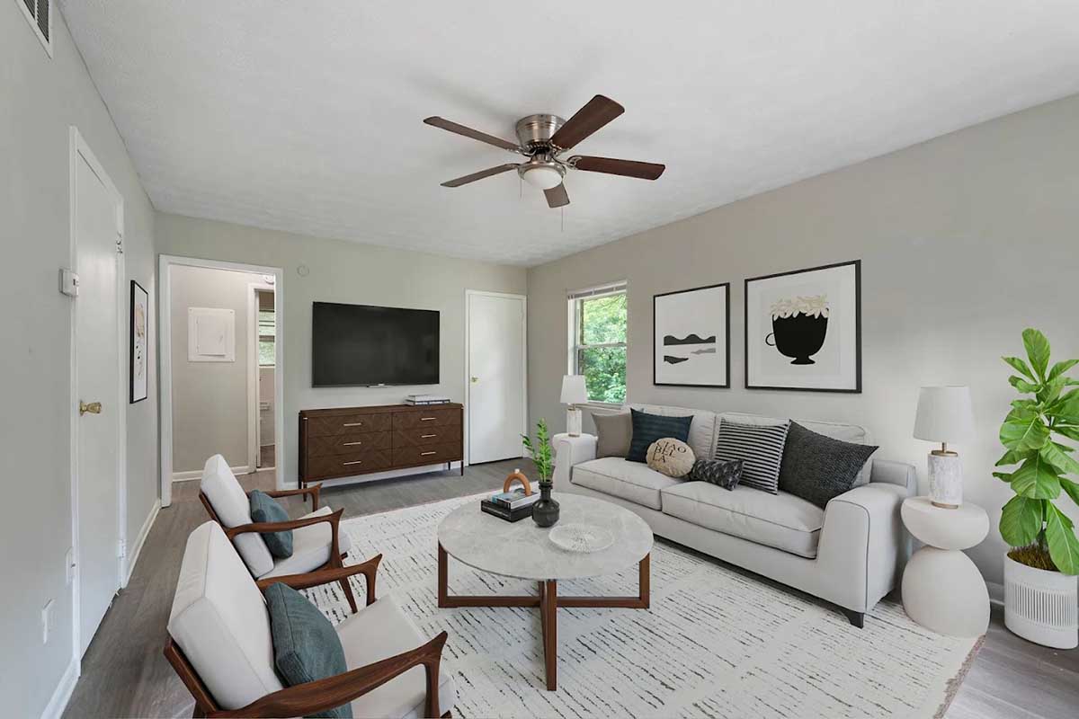 A cozy living room featuring a light gray color scheme. The space includes a comfortable white sofa with black and gray decorative pillows, two wooden chairs, a round coffee table, and a flat-screen TV mounted on the wall. A large window lets in natural light, and there's a potted plant in the corner. Decorative artwork adorns the walls.