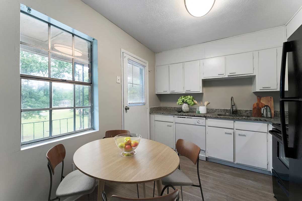 A modern kitchen featuring a round wooden table with four chairs, a bowl of colorful fruit on the table, and white cabinets. Natural light enters through a window, complemented by a black refrigerator and a sink area with granite countertops. The walls are painted light beige.