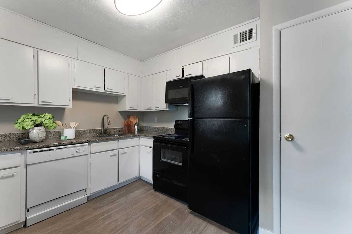 A modern kitchen featuring white cabinets, a black refrigerator, and matching black appliances. The countertop has decorative items, including a small plant. The floor is light-colored laminate, and the space is well-lit with a ceiling light. The kitchen design emphasizes functionality and a clean aesthetic.