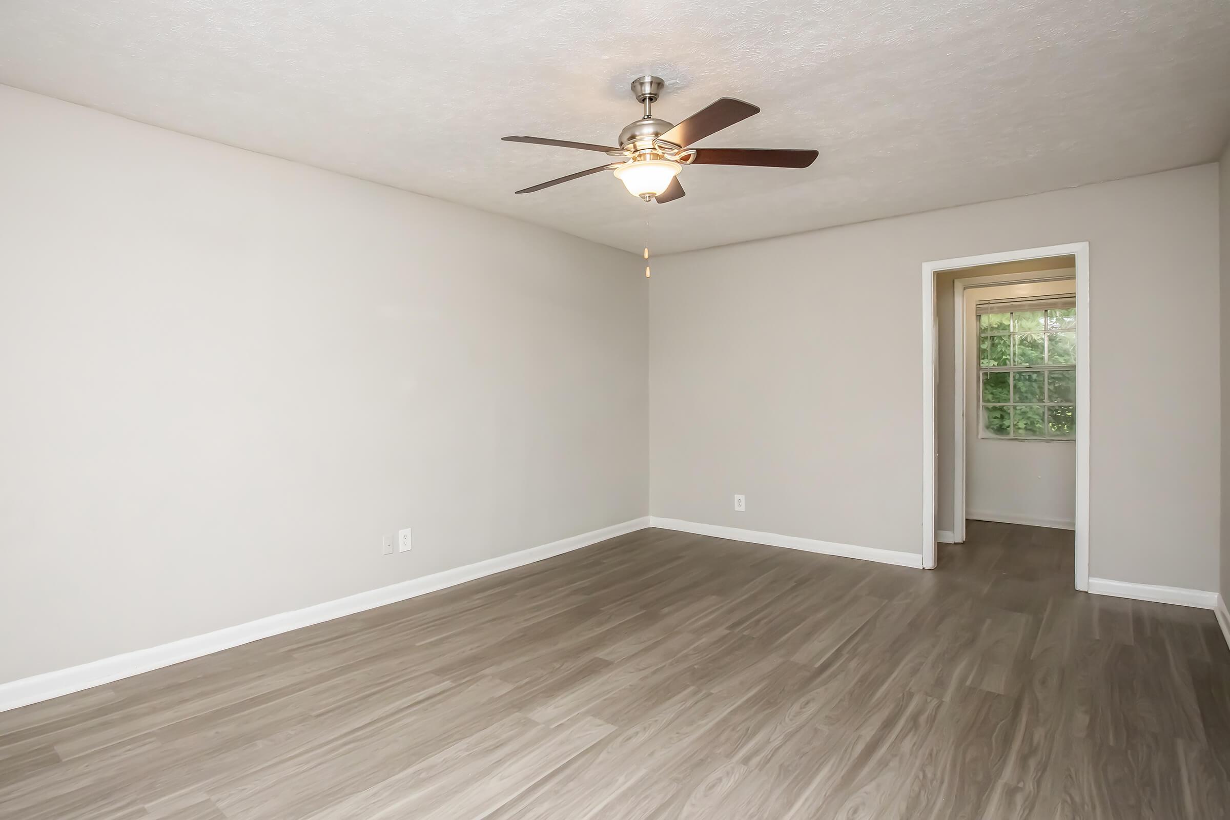A spacious, empty room with light-colored walls and a ceiling fan. The floor is made of light wood panels, and there is an open doorway leading to another room with a window visible in the background. The room is well-lit and has a clean, modern aesthetic.