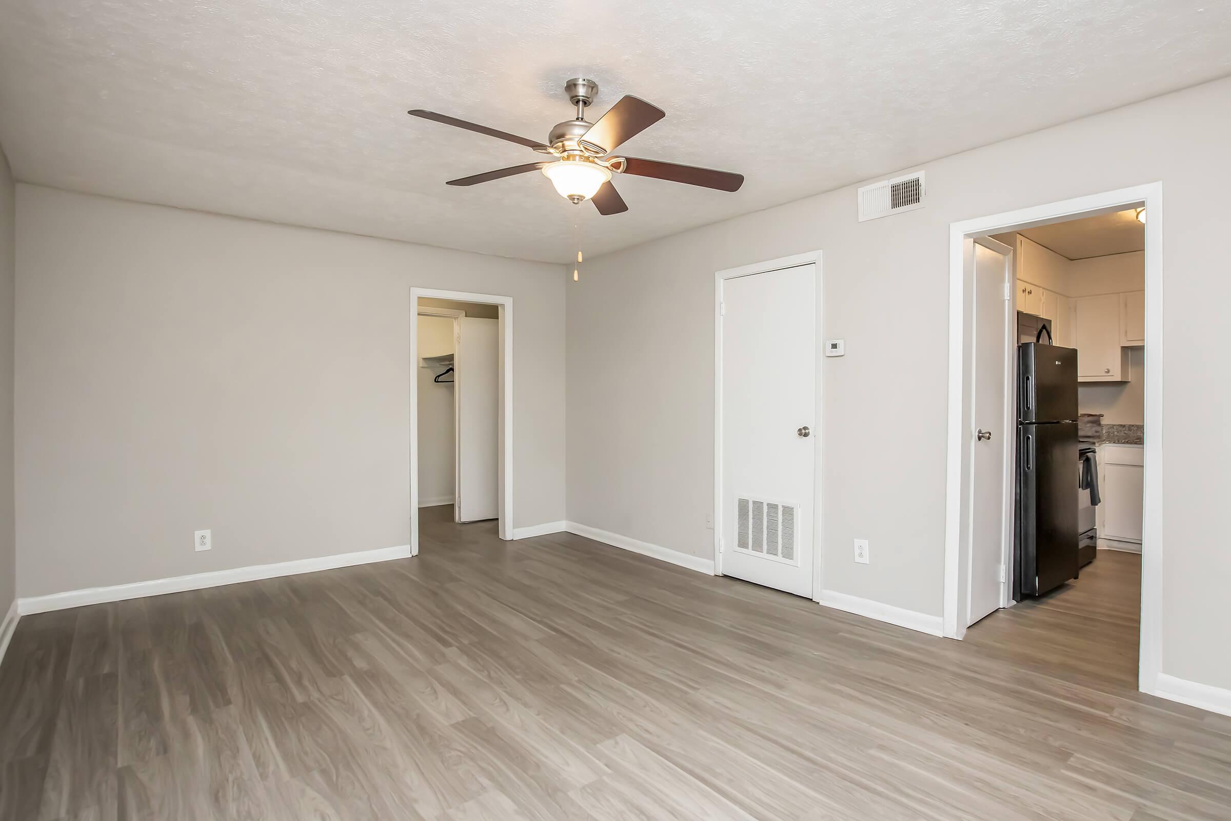 A spacious, empty living room with a ceiling fan, light-colored walls, and laminate flooring. There are two doorways leading to other rooms, one of which appears to lead to a kitchen area with a refrigerator visible. The overall ambiance conveys a clean and open living space.