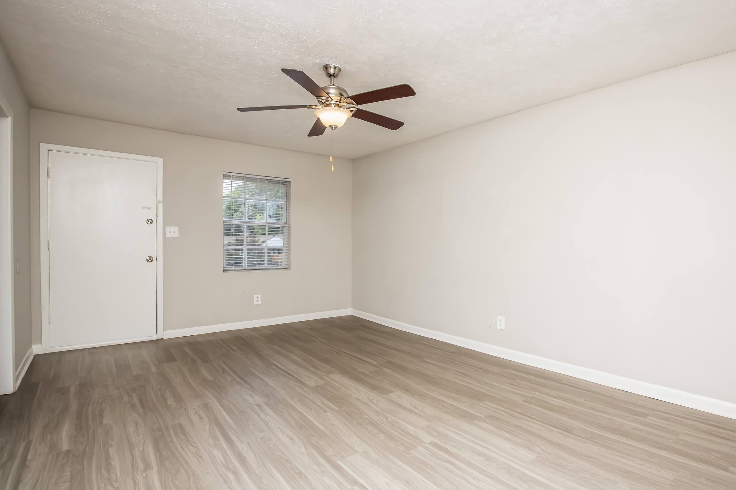 A light-colored living room featuring a ceiling fan, a small window, and a white door. The floor is covered with light wood-like laminate flooring. The walls are a soft neutral color, creating a bright and airy atmosphere. The space is empty, providing a blank canvas for decorating or furnishing.