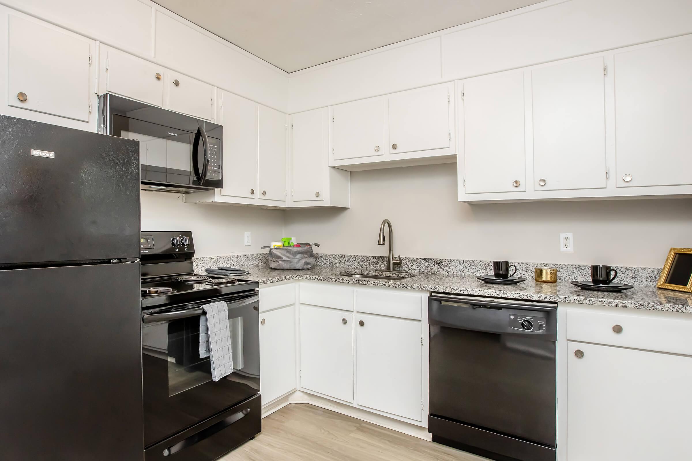Modern kitchen featuring white cabinets, stainless steel appliances including a microwave and dishwasher, a black refrigerator, and a granite countertop. The space is well-lit and organized, with a sink and utensils visible.
