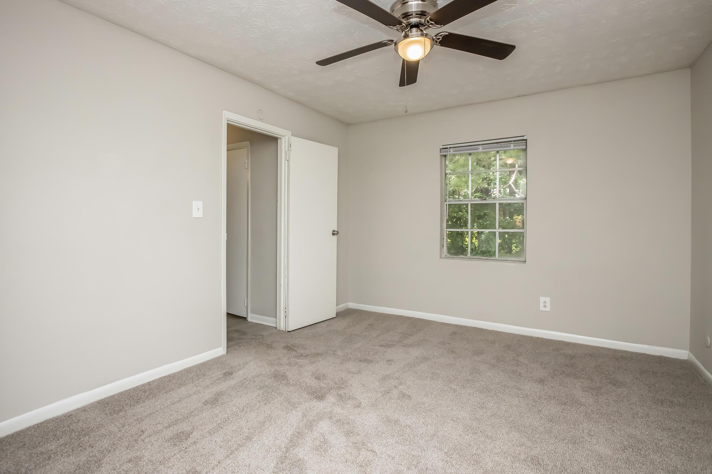 A spacious, empty bedroom featuring light gray walls, carpet flooring, a ceiling fan with a light fixture, and a window allowing natural light. A closed door leads to another room, while the walls are bare, suggesting potential for personalization and decoration.