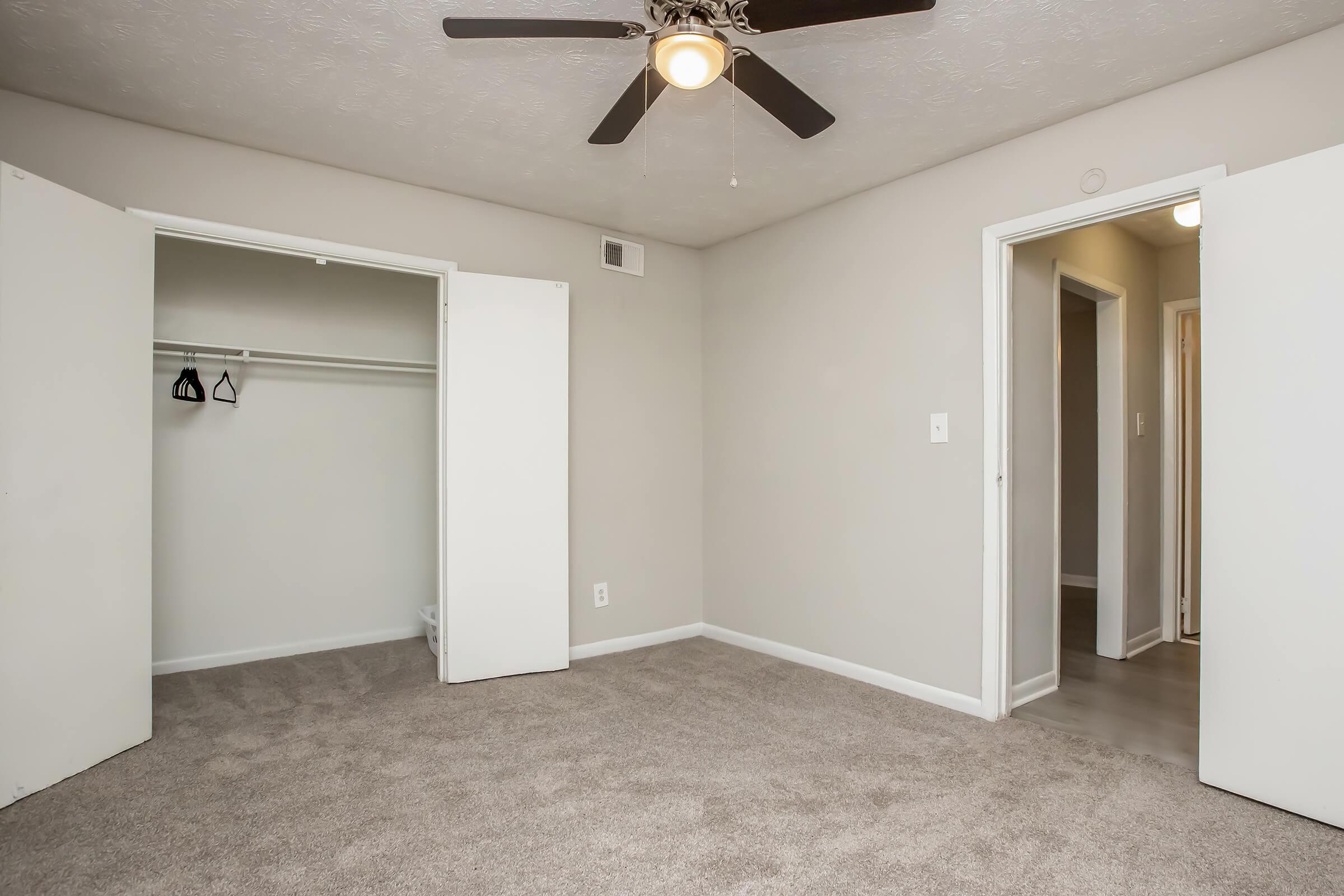 A spacious room with light gray walls and a ceiling fan. There are two open white doors leading to a closet and an adjacent room. The flooring is covered with light-colored carpet, creating a clean and inviting atmosphere. Natural light may enter from the doorway on the right.