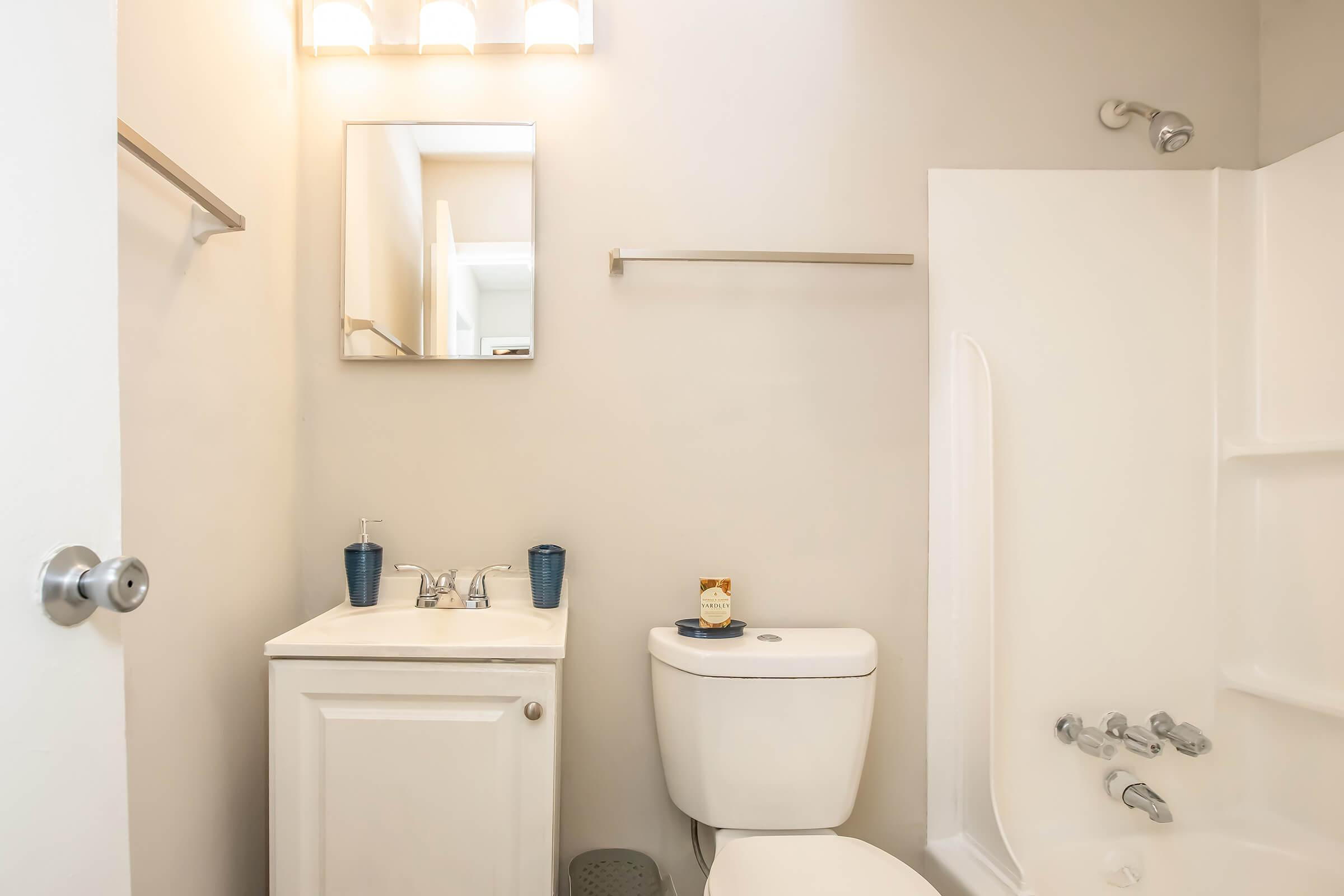 A small bathroom featuring a white toilet, a sink with a modern faucet, a mirror above the sink, and a bathtub with a shower. The walls are a light neutral color, and there are two blue cups on the sink. A towel rack is mounted above the toilet.