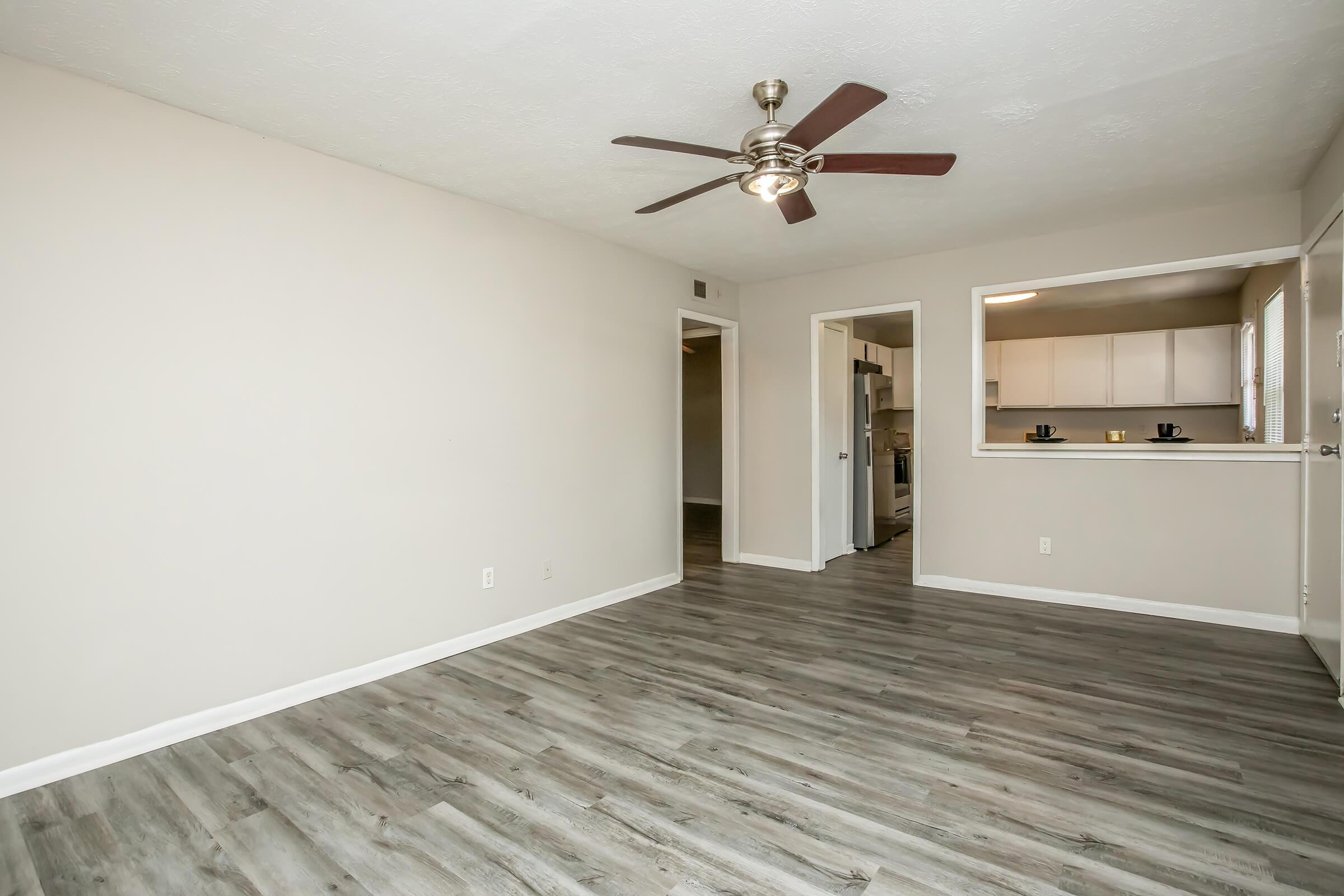 Empty living room featuring light-colored walls, a ceiling fan, and modern laminate flooring. A doorway leads to another room, and an open passageway connects to a kitchen area visible in the background. Natural light brightens the space, creating a clean and airy atmosphere.