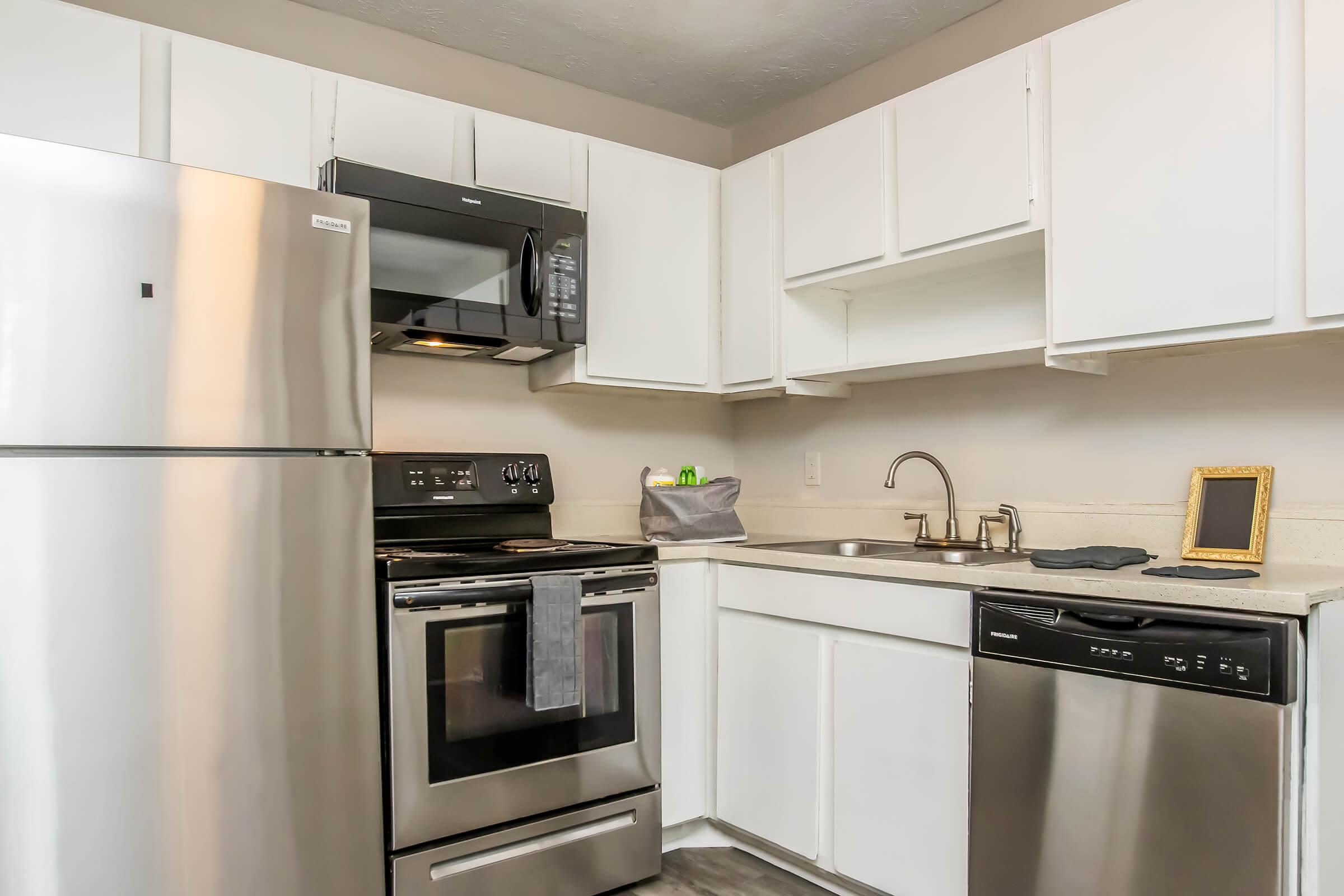 Modern kitchen featuring stainless steel appliances including a refrigerator, microwave, oven, and dishwasher. The cabinetry is white with a light countertop. There is a sink with a faucet and a small decorative mirror on the counter, along with minimal kitchen accessories. The space is well-lit and organized.