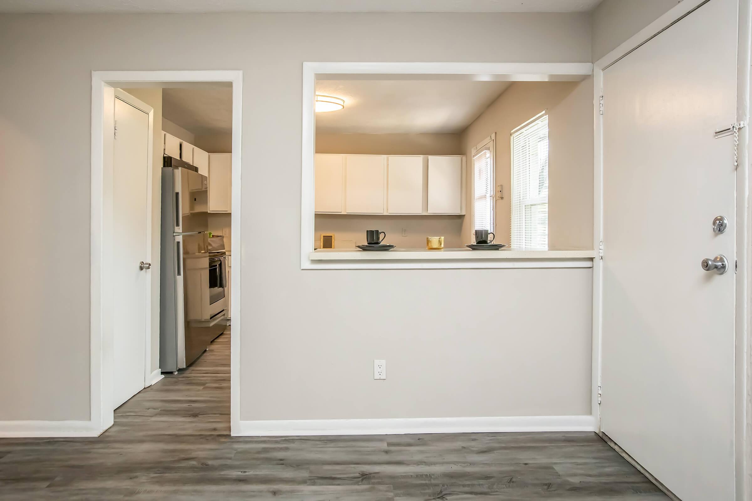 Interior view of a modern apartment, featuring a small entryway with a door leading outside. A kitchen is visible through a pass-through window, showcasing white cabinets and appliances. The flooring is a light wood finish, and the walls are painted a neutral gray. Two black cups are placed on a countertop.
