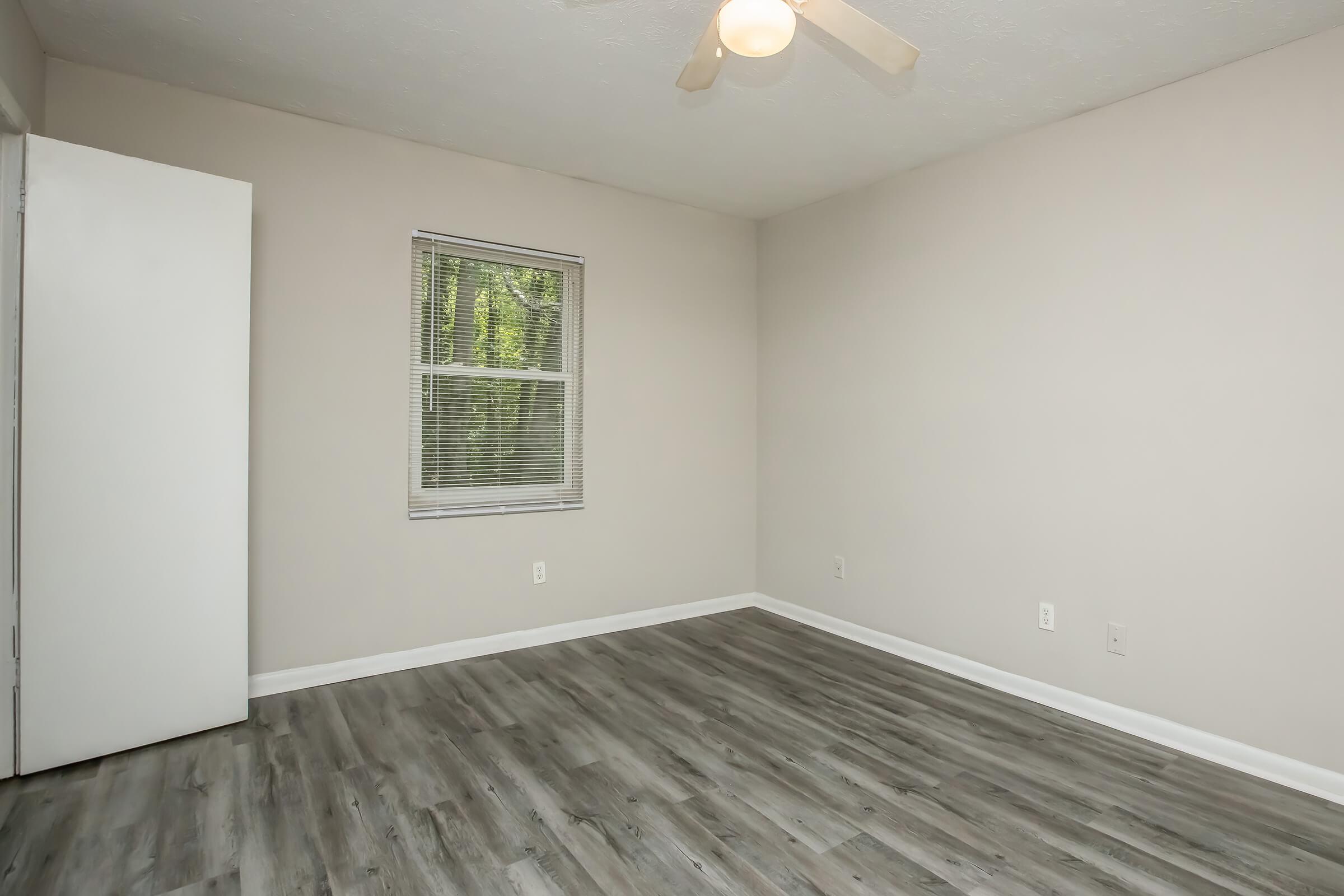 A spacious, empty room with light-colored walls and a wooden floor. There is a ceiling fan and a single window with blinds, allowing natural light to enter. A closed white door is visible on the left side. The room has a clean and minimalist appearance, suitable for various uses.