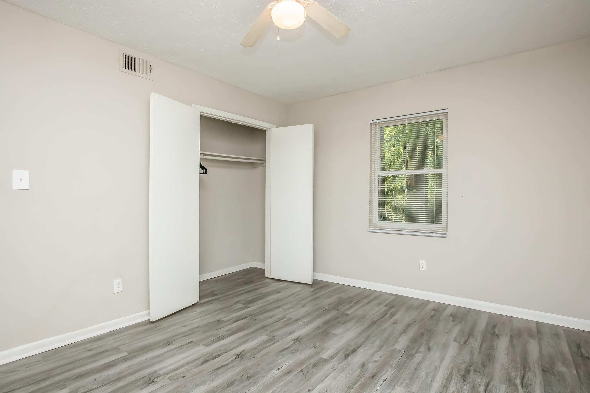 A spacious empty room with light gray walls and a wooden floor. There's a ceiling fan with a light fixture, a window with blinds letting in natural light, and an open closet with white sliding doors. The overall vibe is bright and uncluttered, making it suitable for various uses.