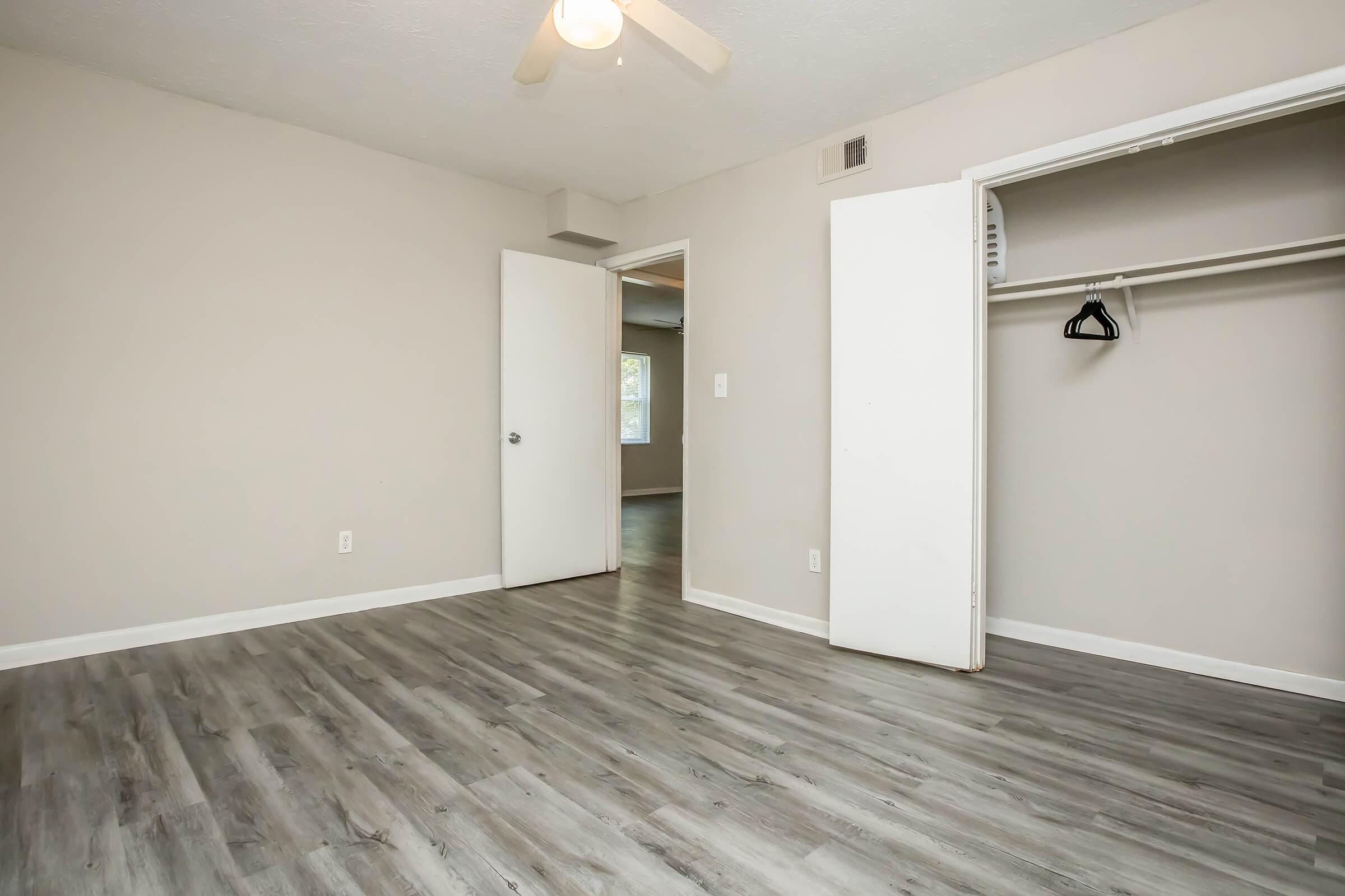 A light-colored room with a ceiling fan and smooth wooden flooring. There is an open closet with a hanger visible, and a doorway leading to another room in the background. The walls are painted a neutral shade, creating a bright and airy atmosphere.
