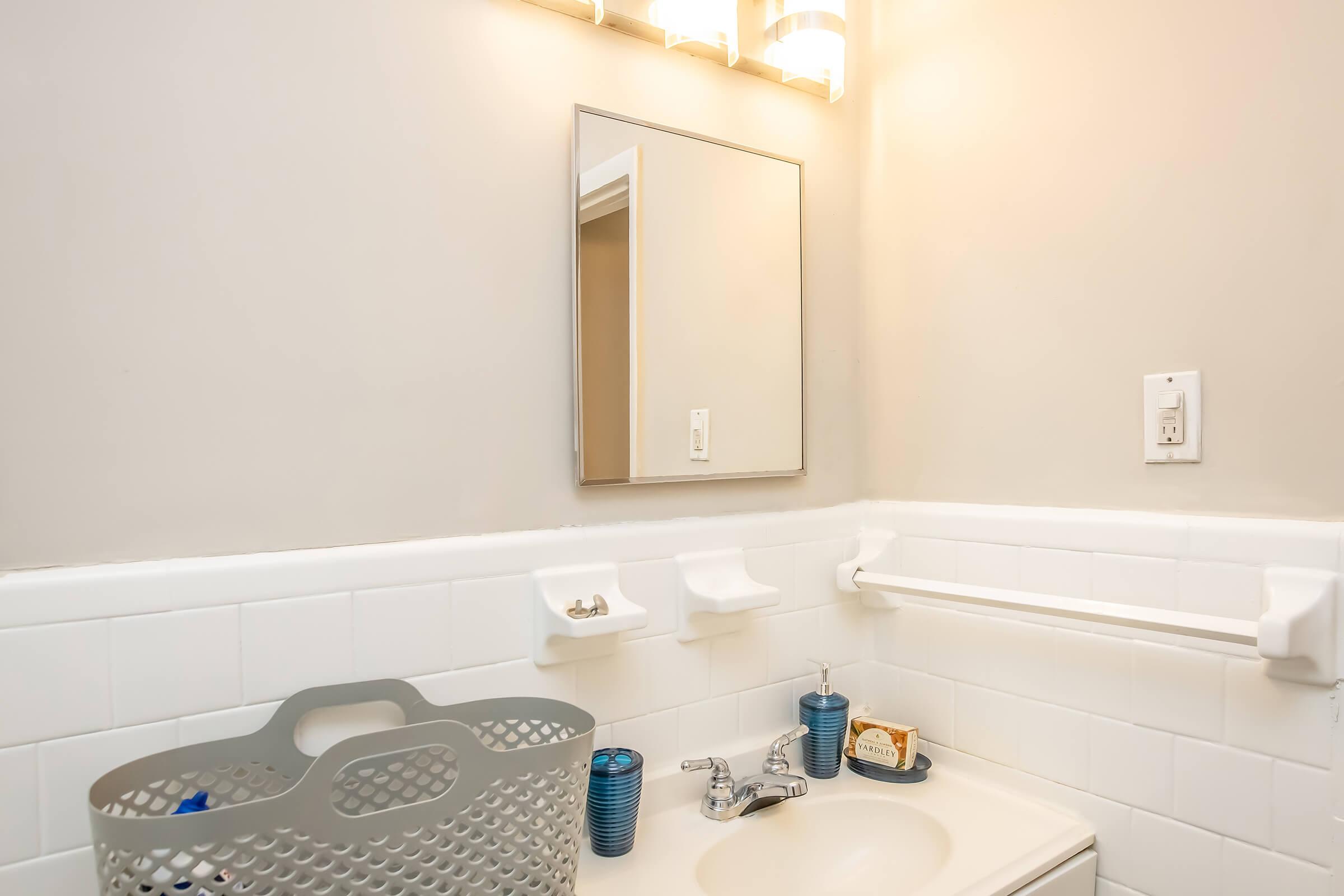 A clean and simple bathroom with a wall-mounted mirror above a sink. The sink has a faucet and is accompanied by a gray basket and a small decorative item. Light fixtures above the mirror provide illumination, and there are white tiled walls with hooks for towels.
